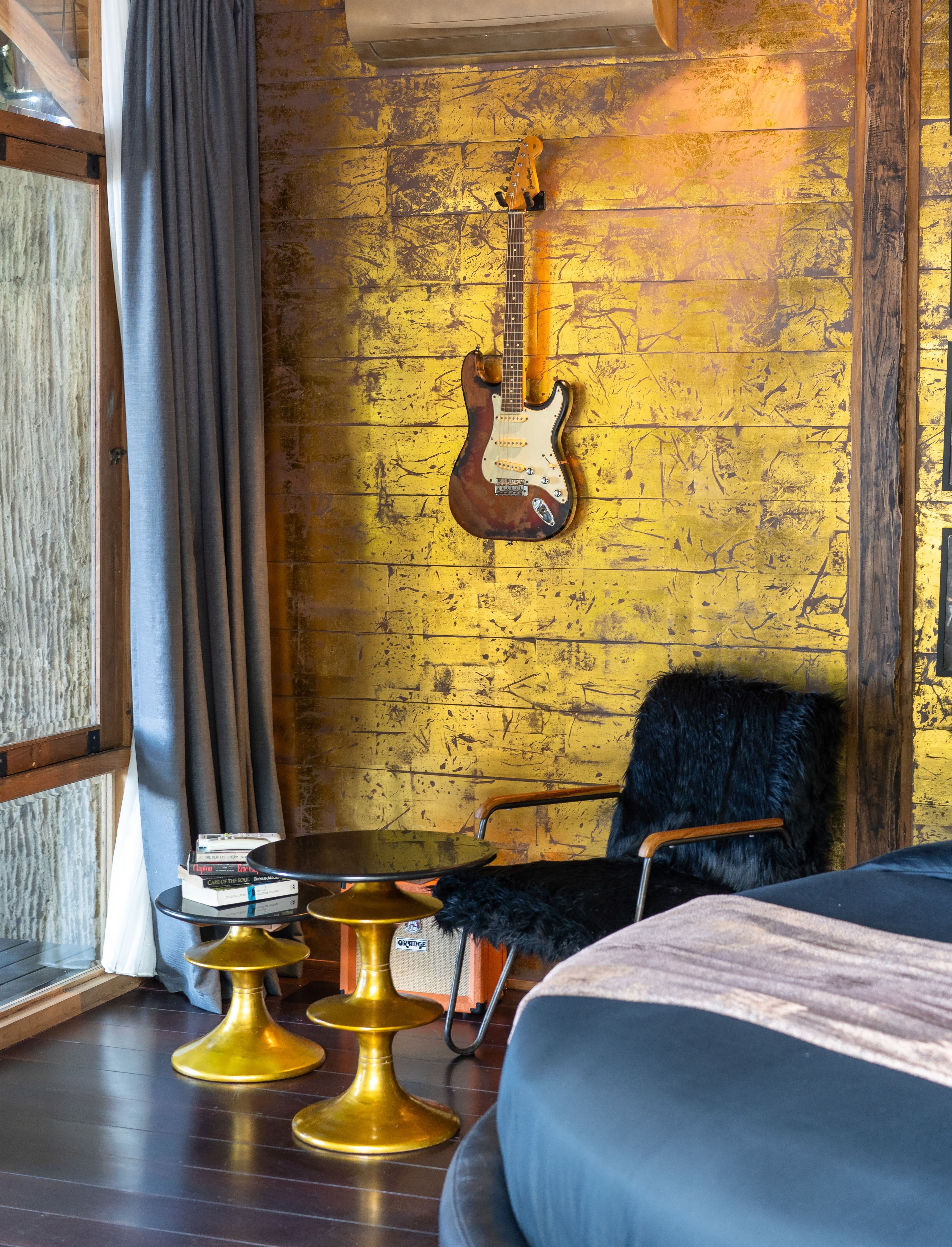A guitar mounted on the yellow brick wall above a fur-draped chair and a stack of books.