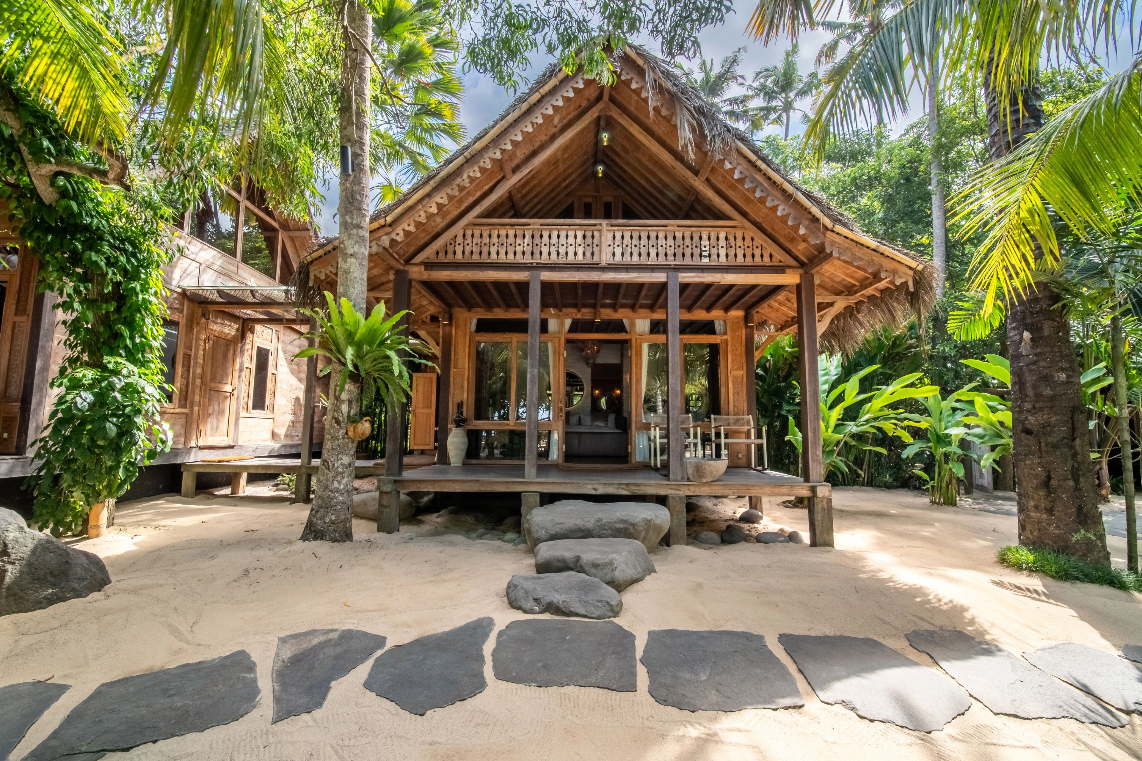 The carved timber façade of the villa across the sand courtyard, stepping stones leading to it.