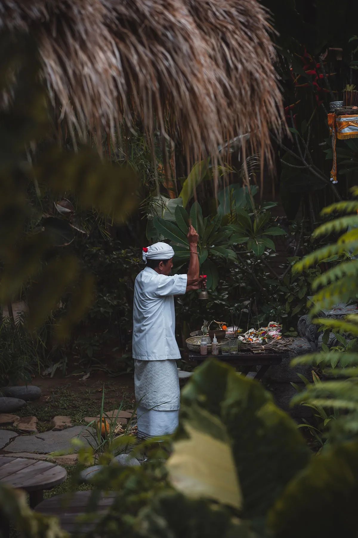 A priest in white performing a blessing over offerings laid out in the garden.