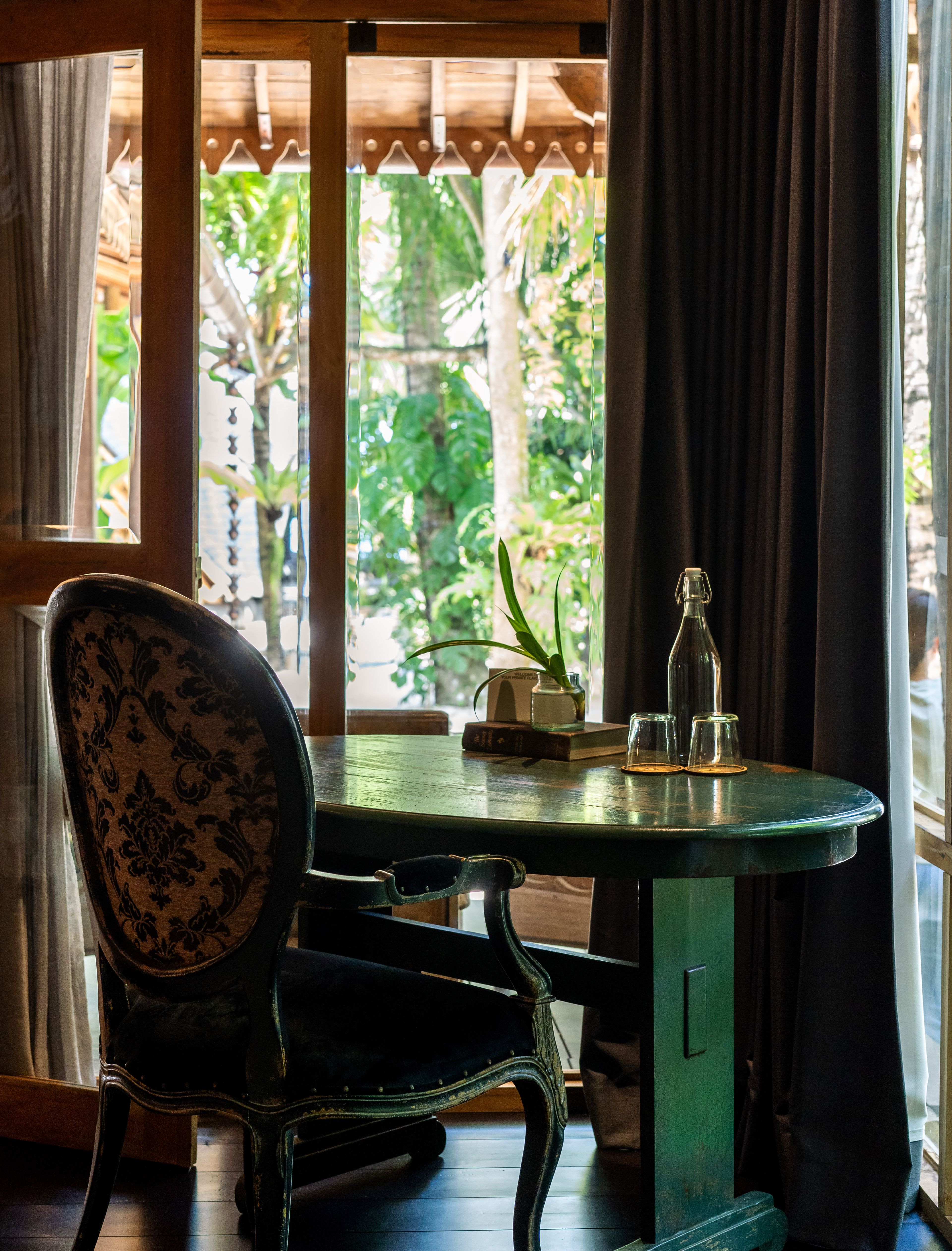A carved chair at a round green table with a bottle and glasses, garden beyond the window.