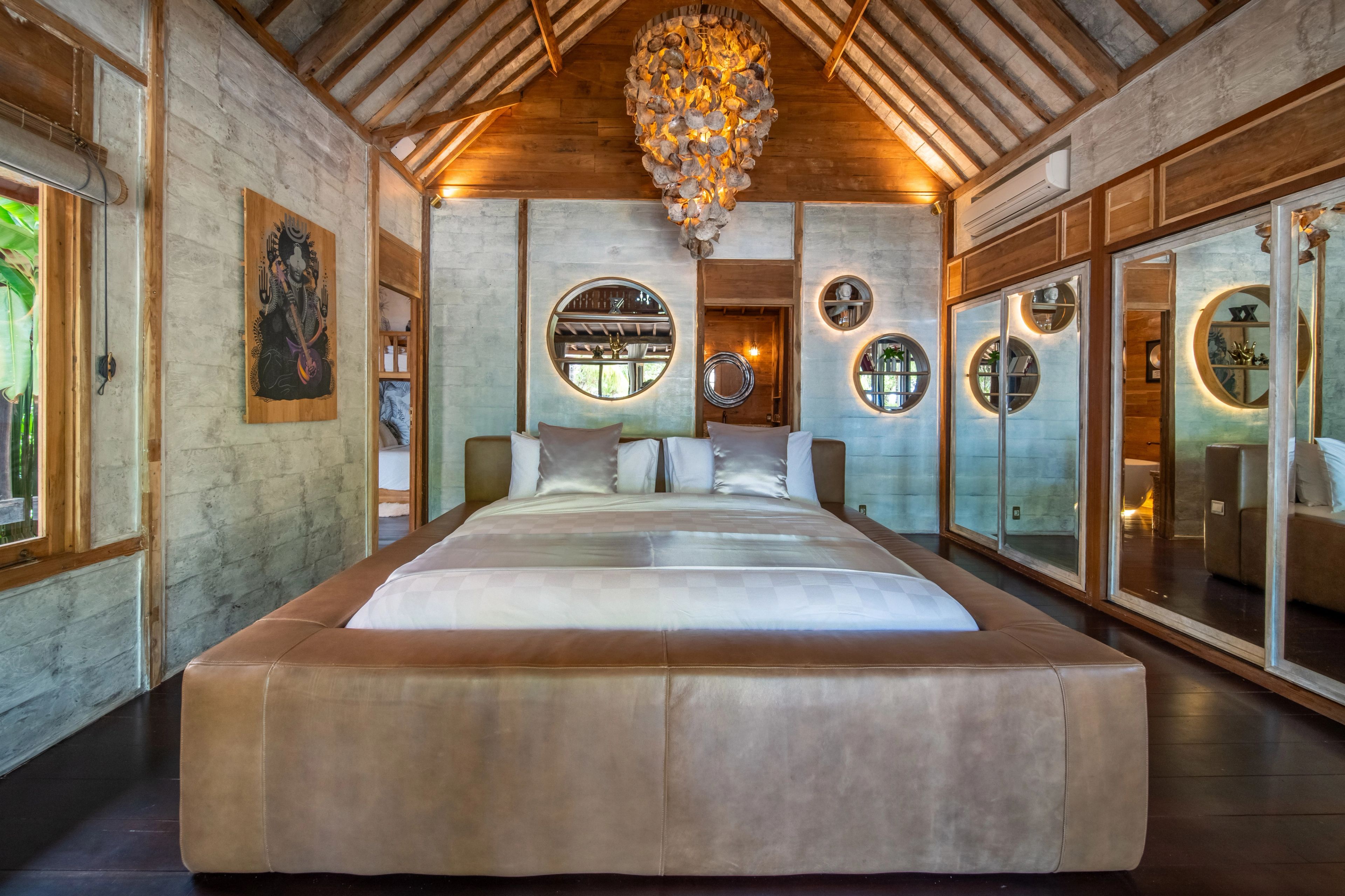 A wide bed with a leather base under the shell chandelier, round mirrors set into the wall alongside.