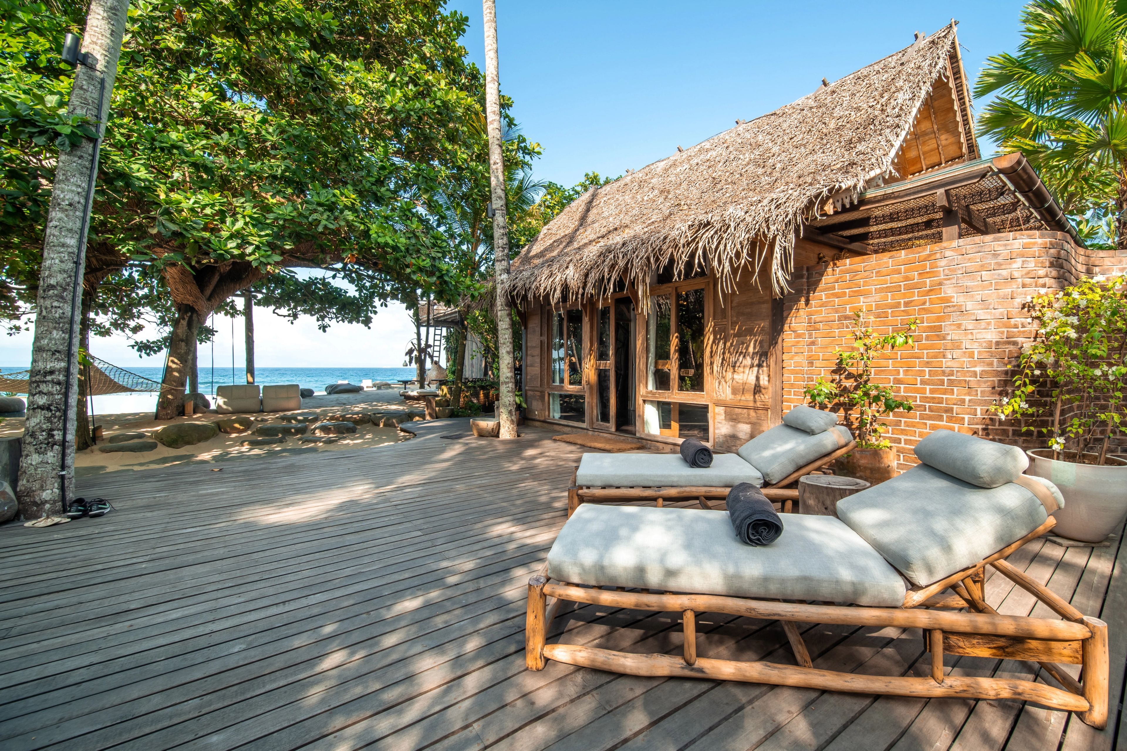 Loungers on the timber deck outside the thatched villa, the beach and sea just beyond the trees.