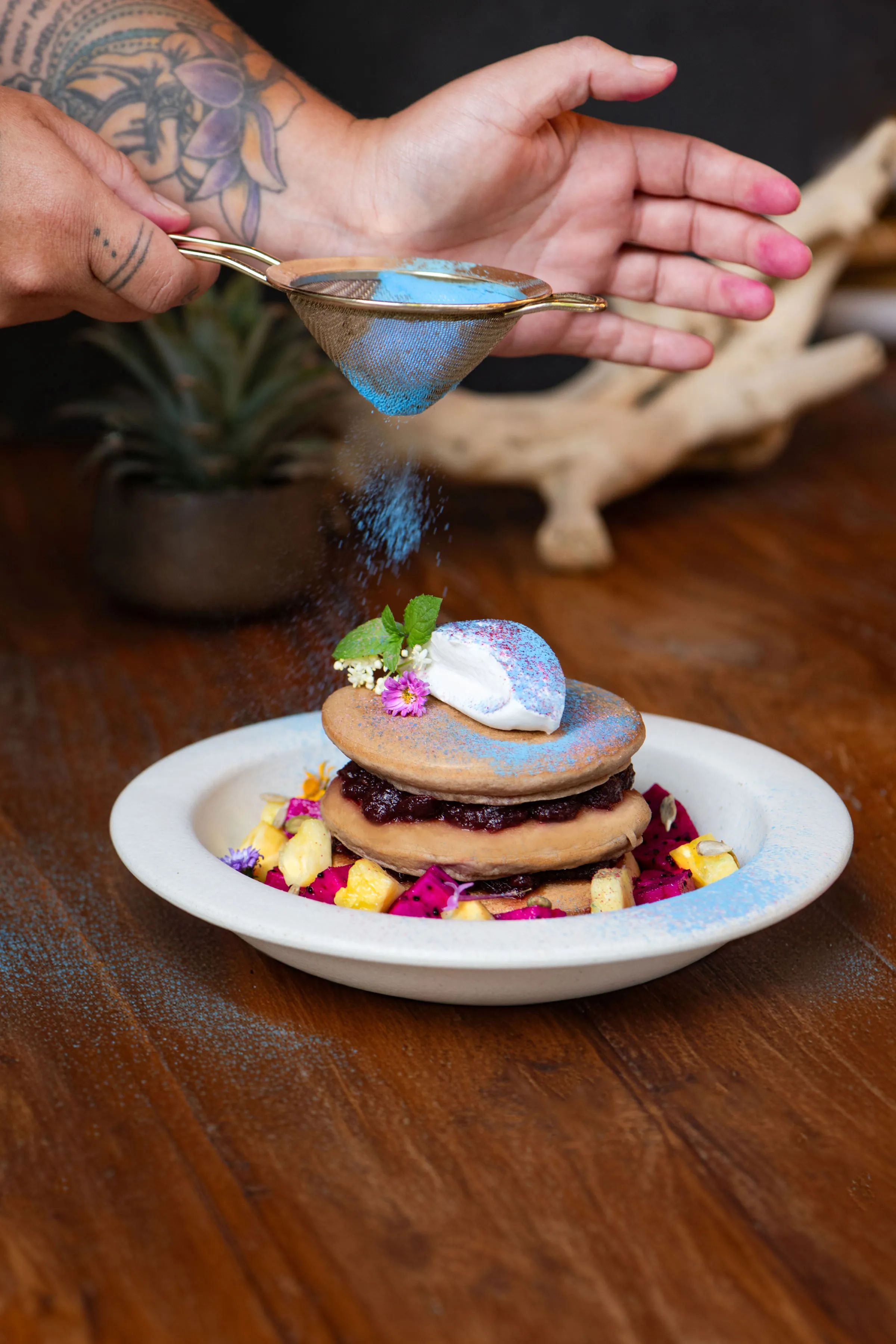 A stack of pancakes layered with compote and cream, dusted with blue powder and scattered with flowers.