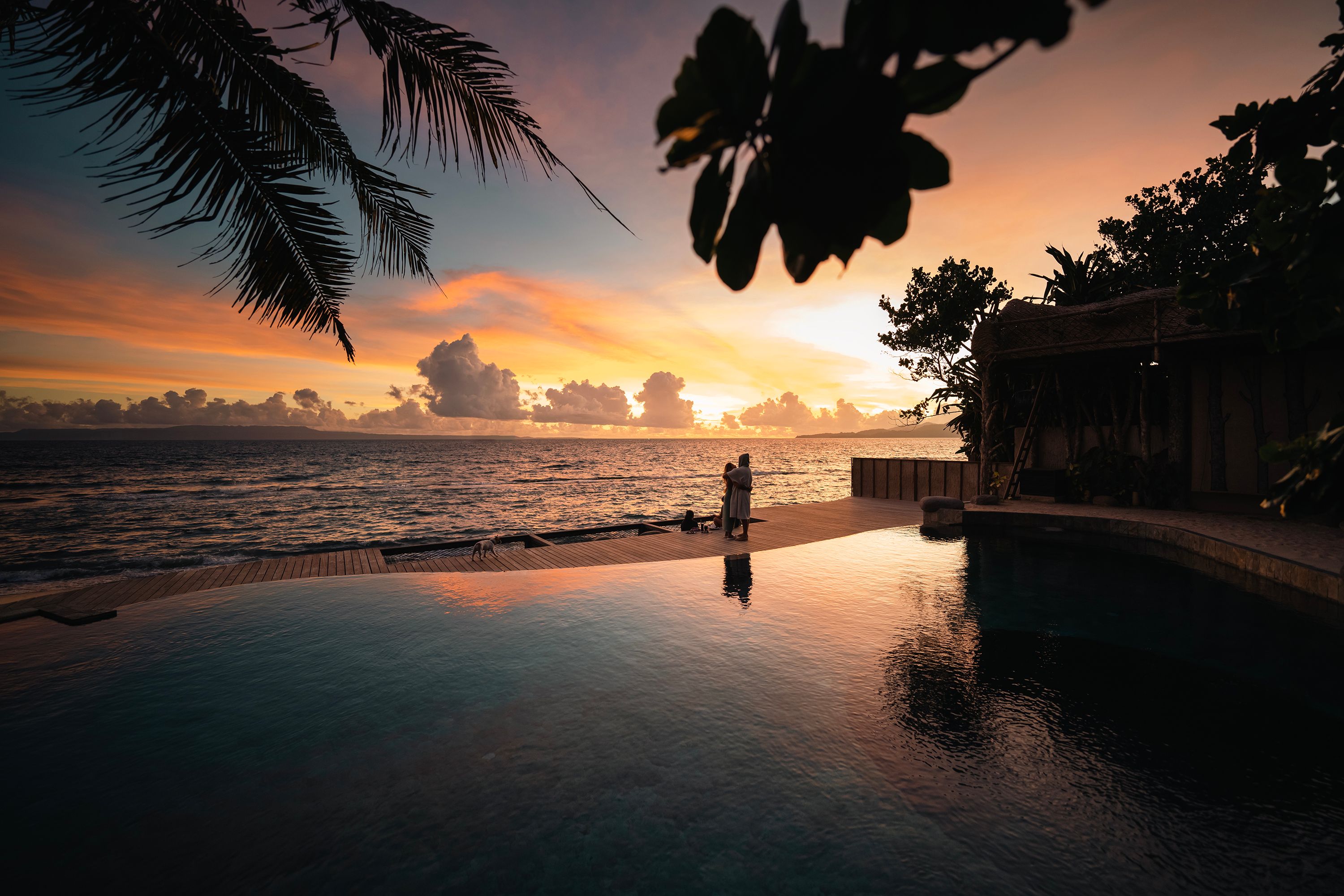 The sun setting beyond the infinity pool, the sky reflected in the still water and palms in silhouette.