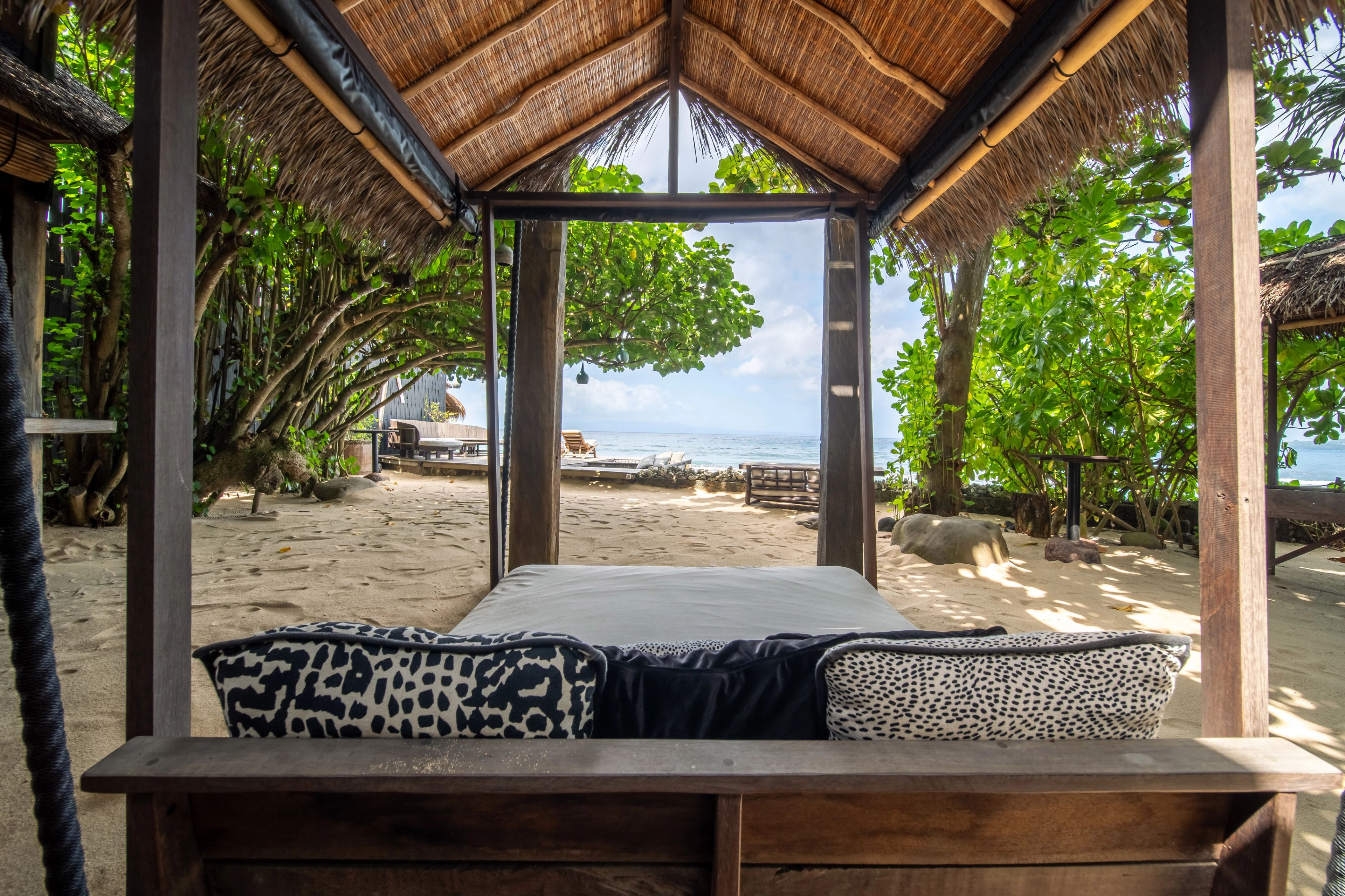 A daybed under a thatched cabana roof, looking down the beach through the trees.