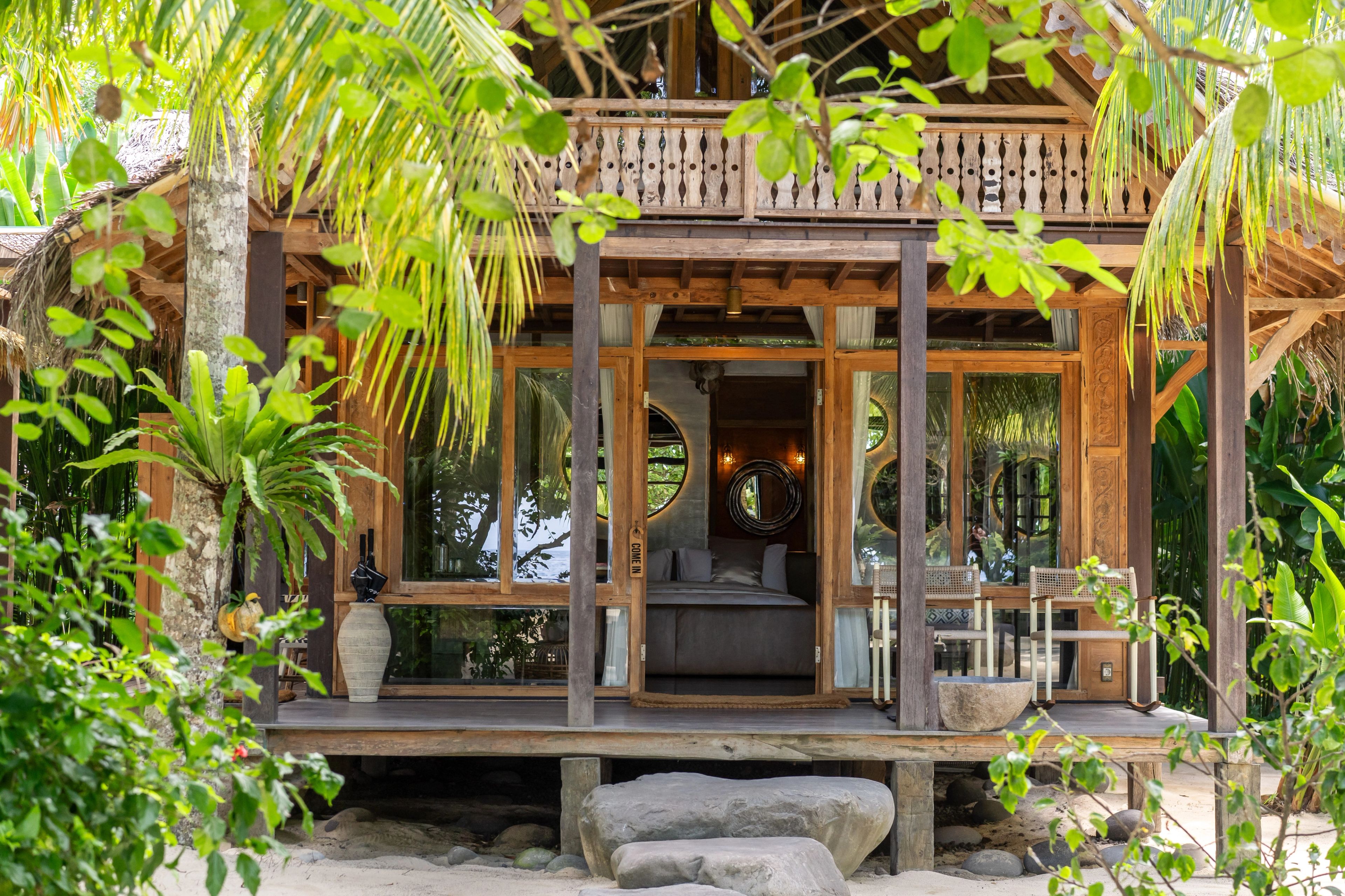 The two-storey timber villa seen through the planting, its balcony above the open doors.