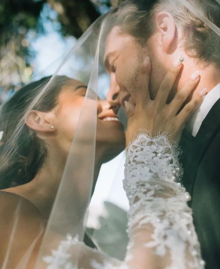 A close view of a couple kissing beneath the bride's veil, her beaded sleeve at his cheek.