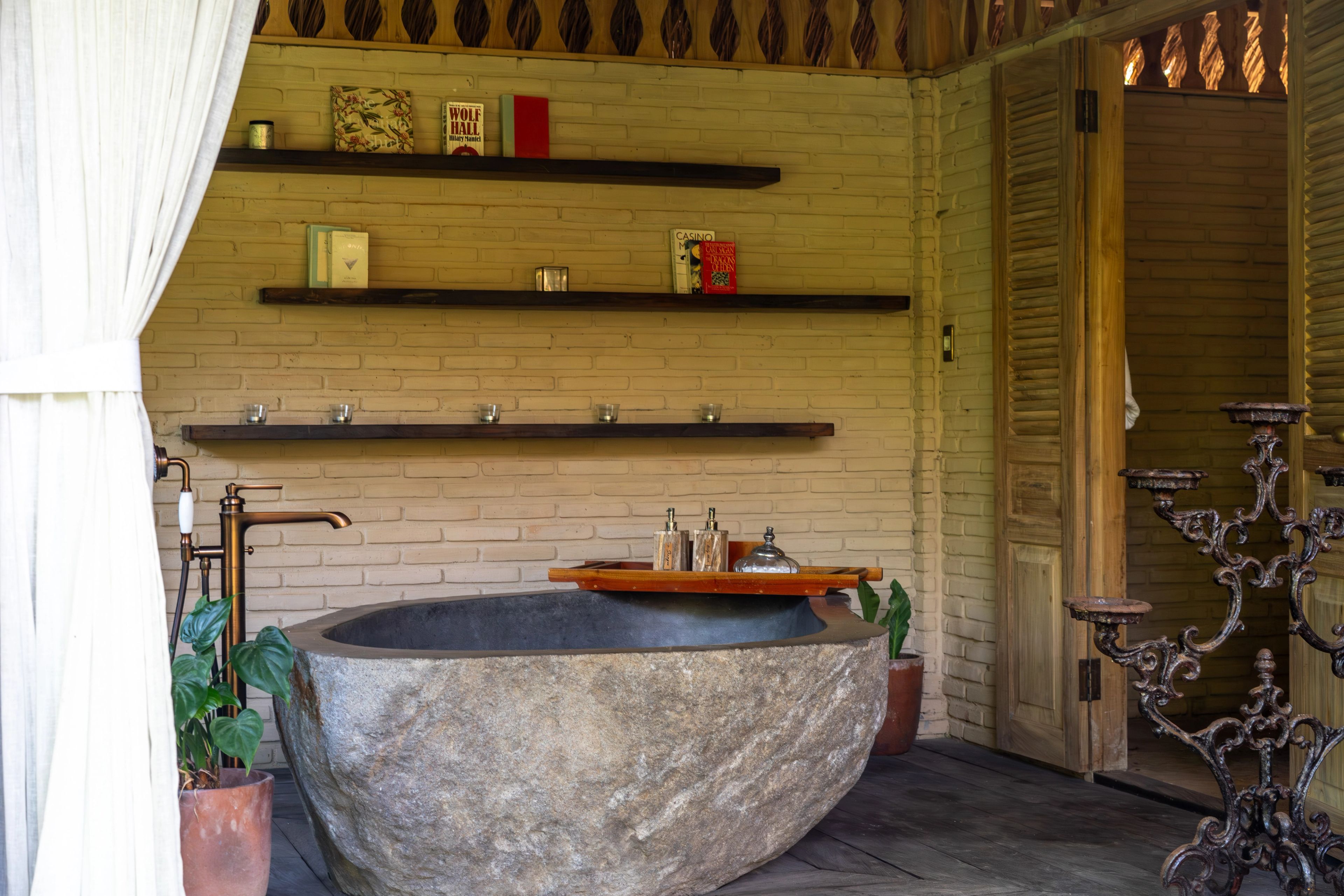 A carved stone bathtub beside a wrought-iron candelabra, with a tray of bottles on a timber shelf.