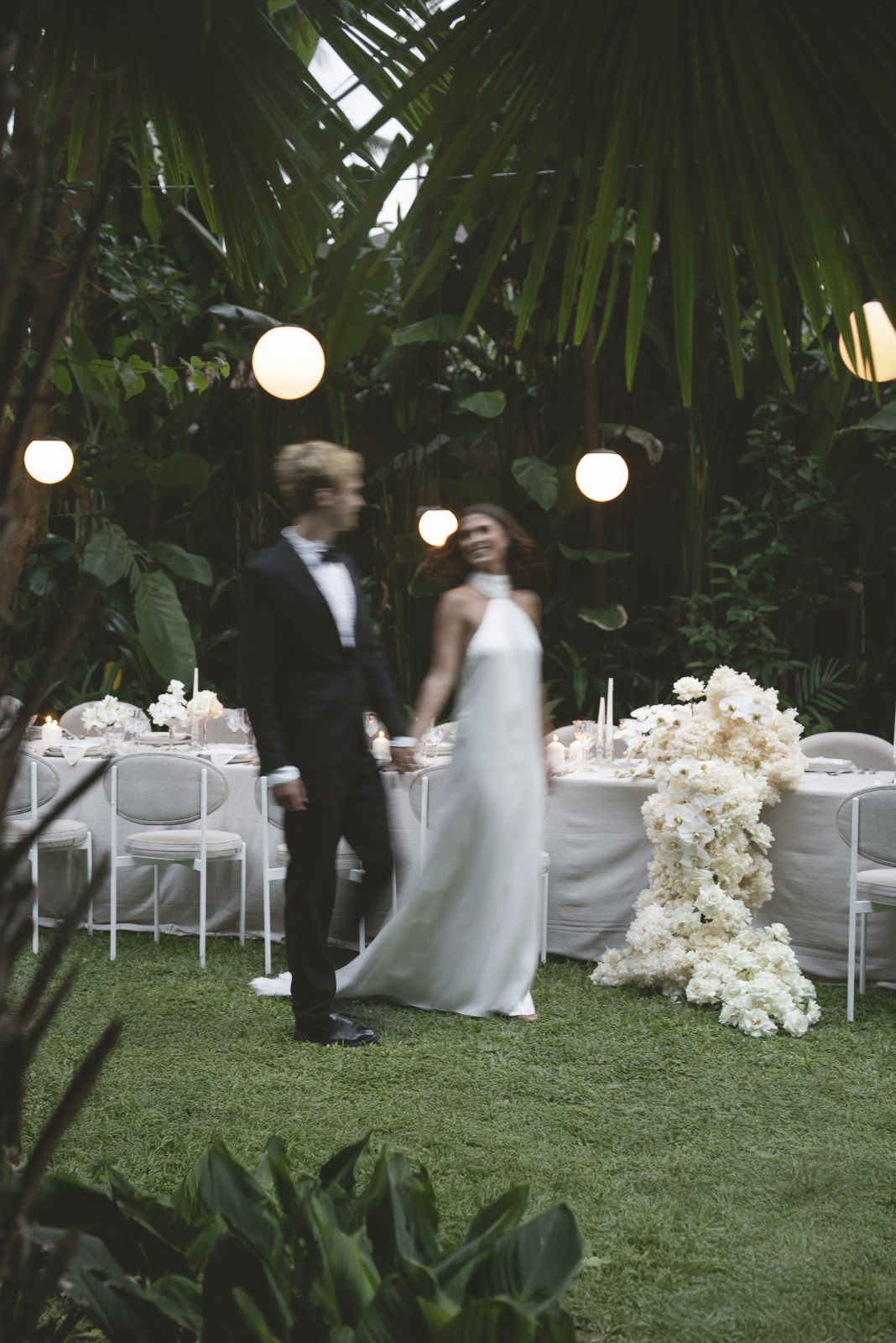 A couple walking past the set table in the garden under strung festoon lights.