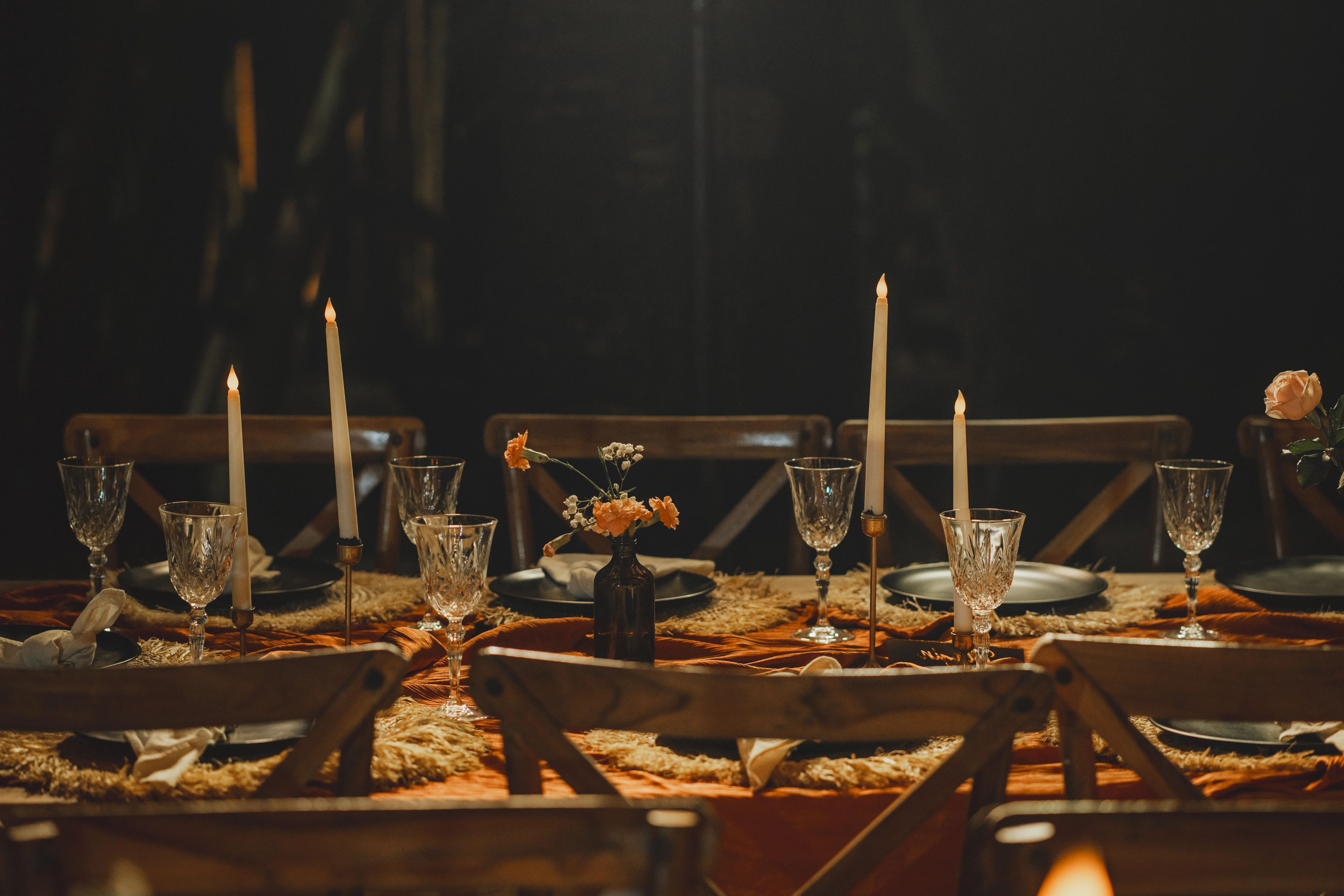 Candles, glassware and small vases running down a table laid for dinner in low light.