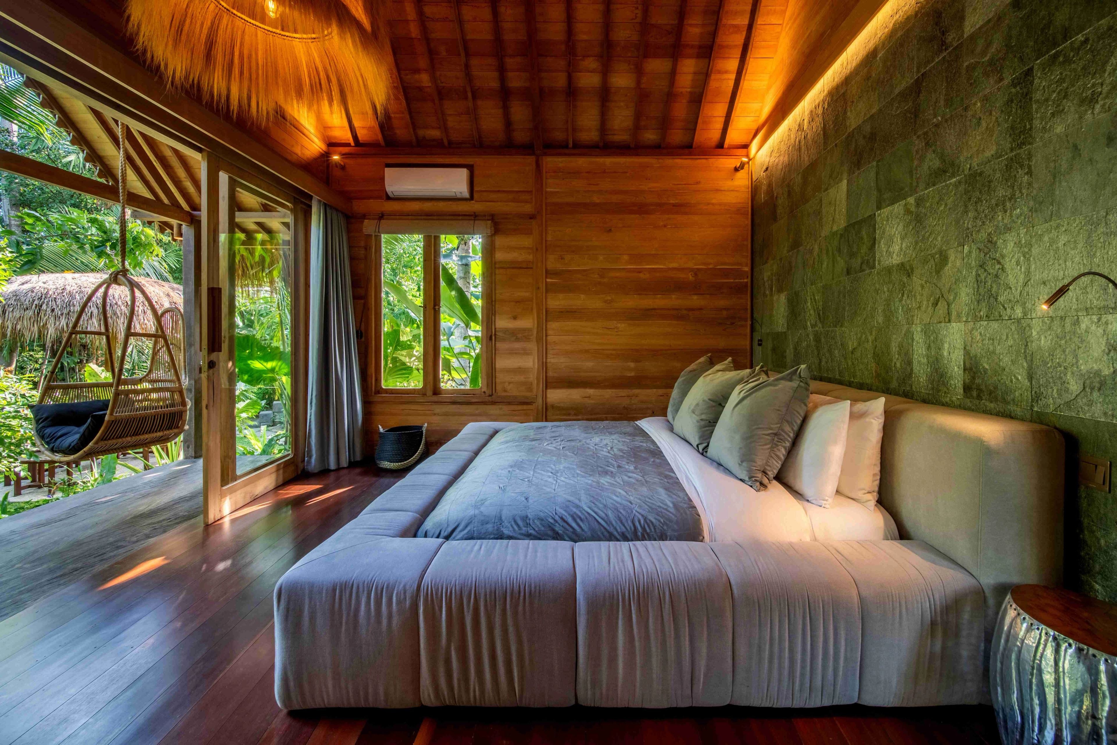 A wide bed under the thatched roof, a hanging chair by the window and a stone wall behind the headboard.