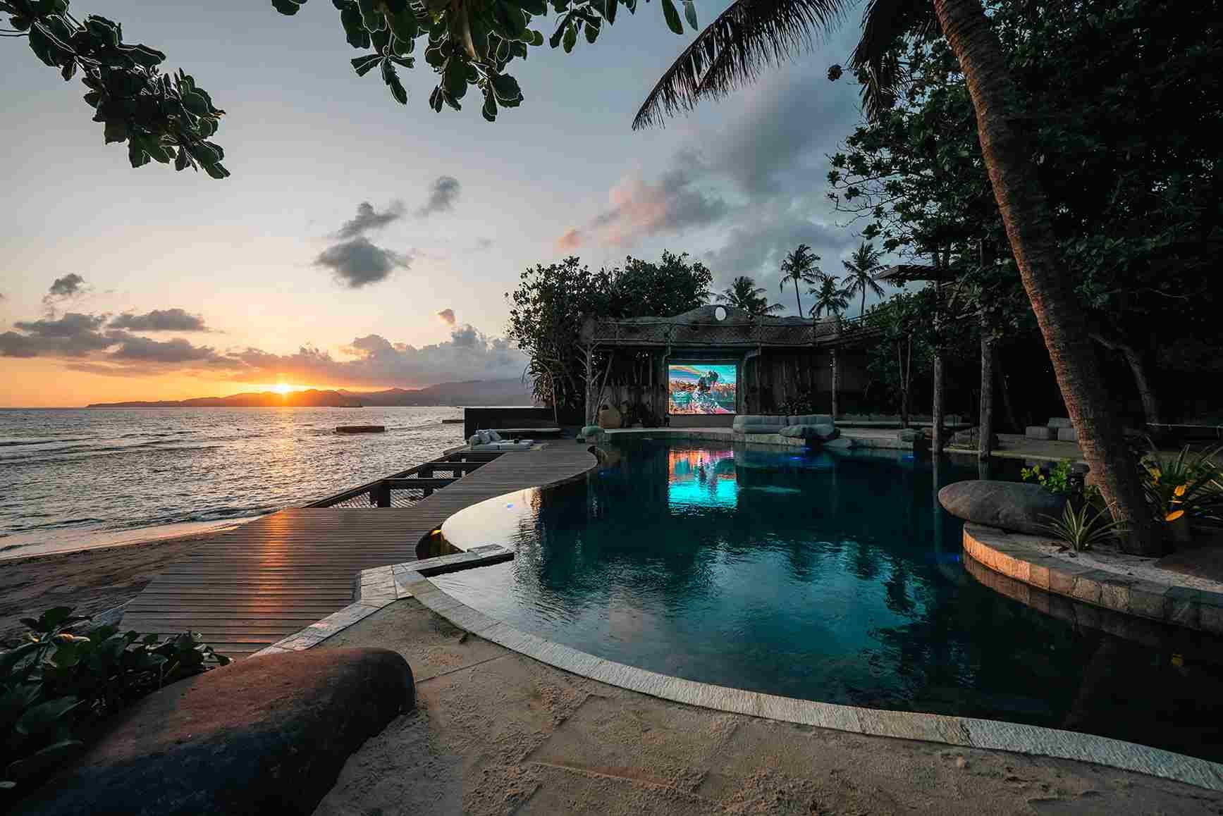 The sun setting over the sea beyond the lit pool and the outdoor screen.