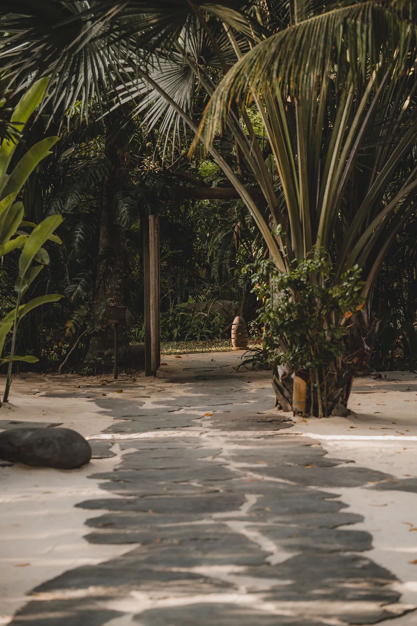 A sand and stone courtyard under a pandanus, enclosed by timber posts and dense planting.