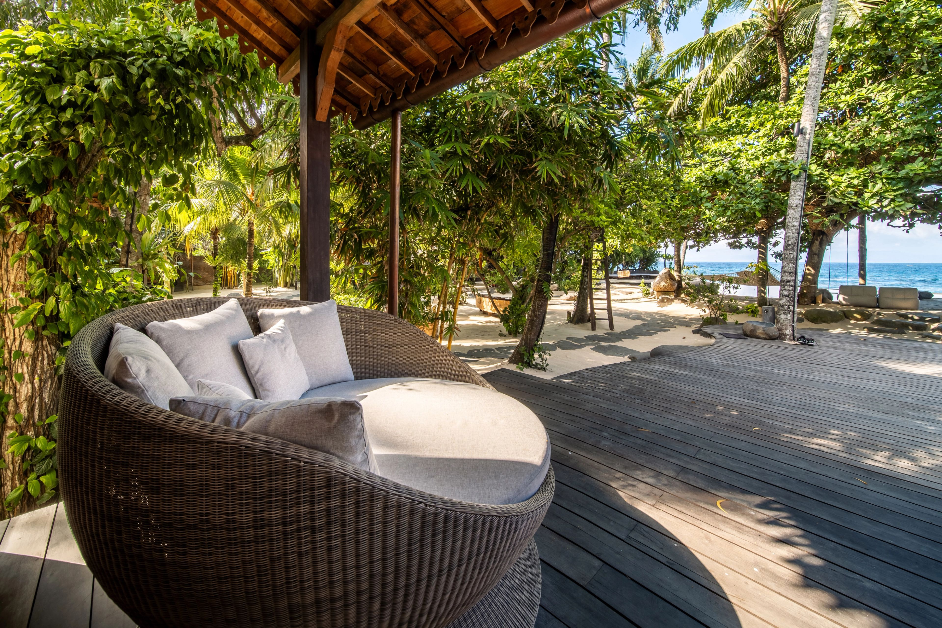 A round woven daybed on the deck under a planted pergola, the sea beyond.