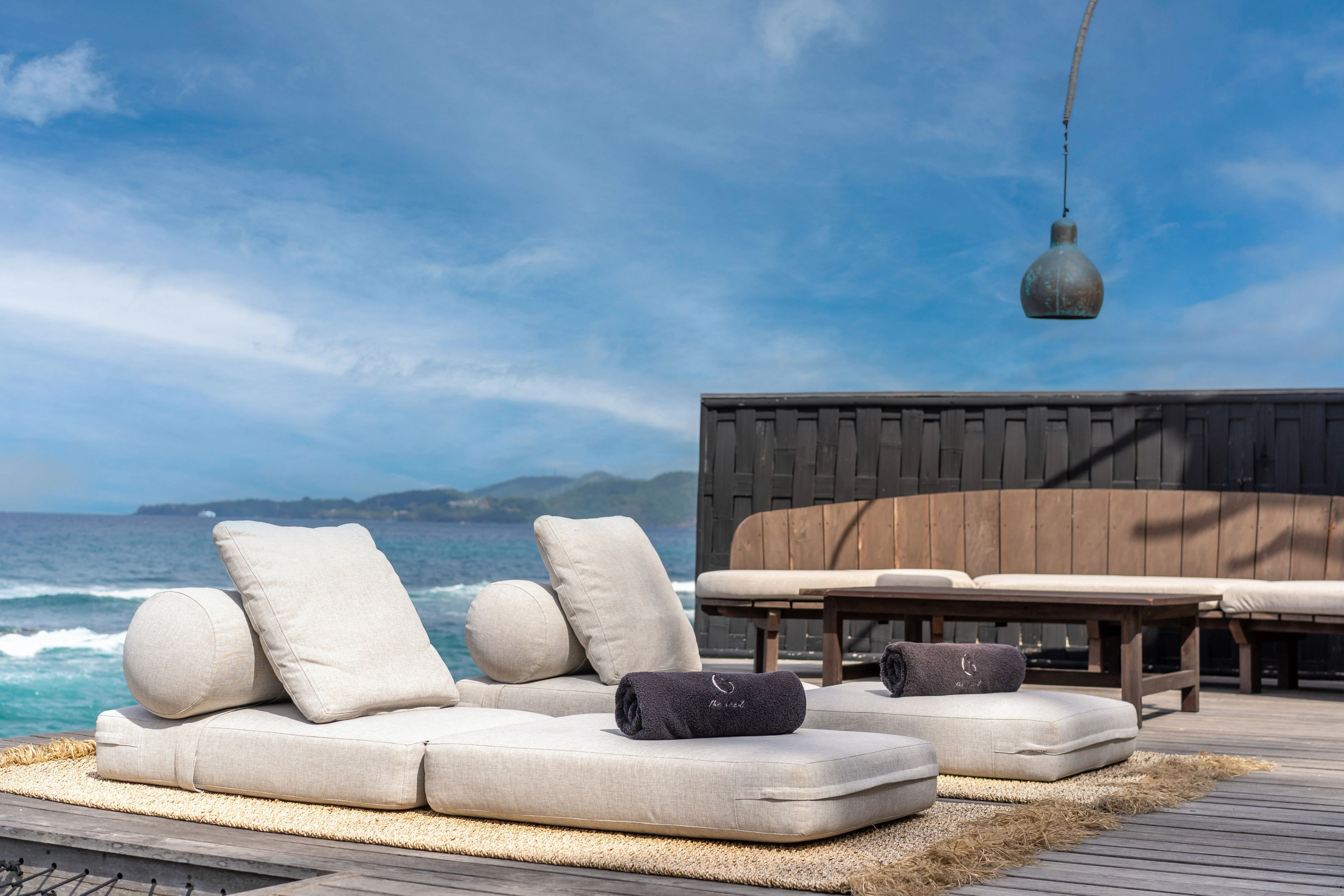 Two daybeds with bolsters and rolled towels on the deck under a hanging lamp, the sea beyond.
