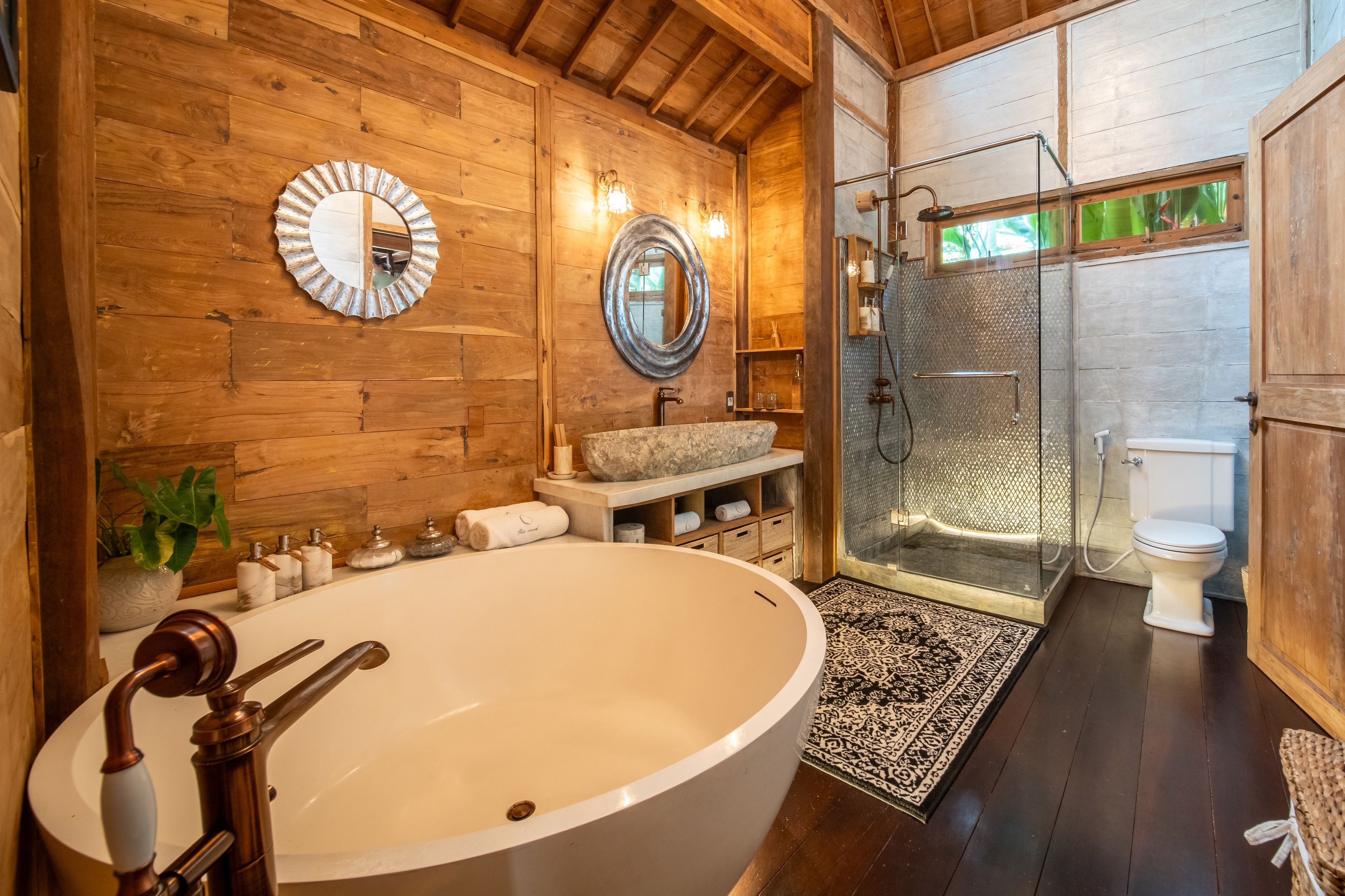 A freestanding oval bath with a stone basin and glass shower behind, mirrors on the timber wall.