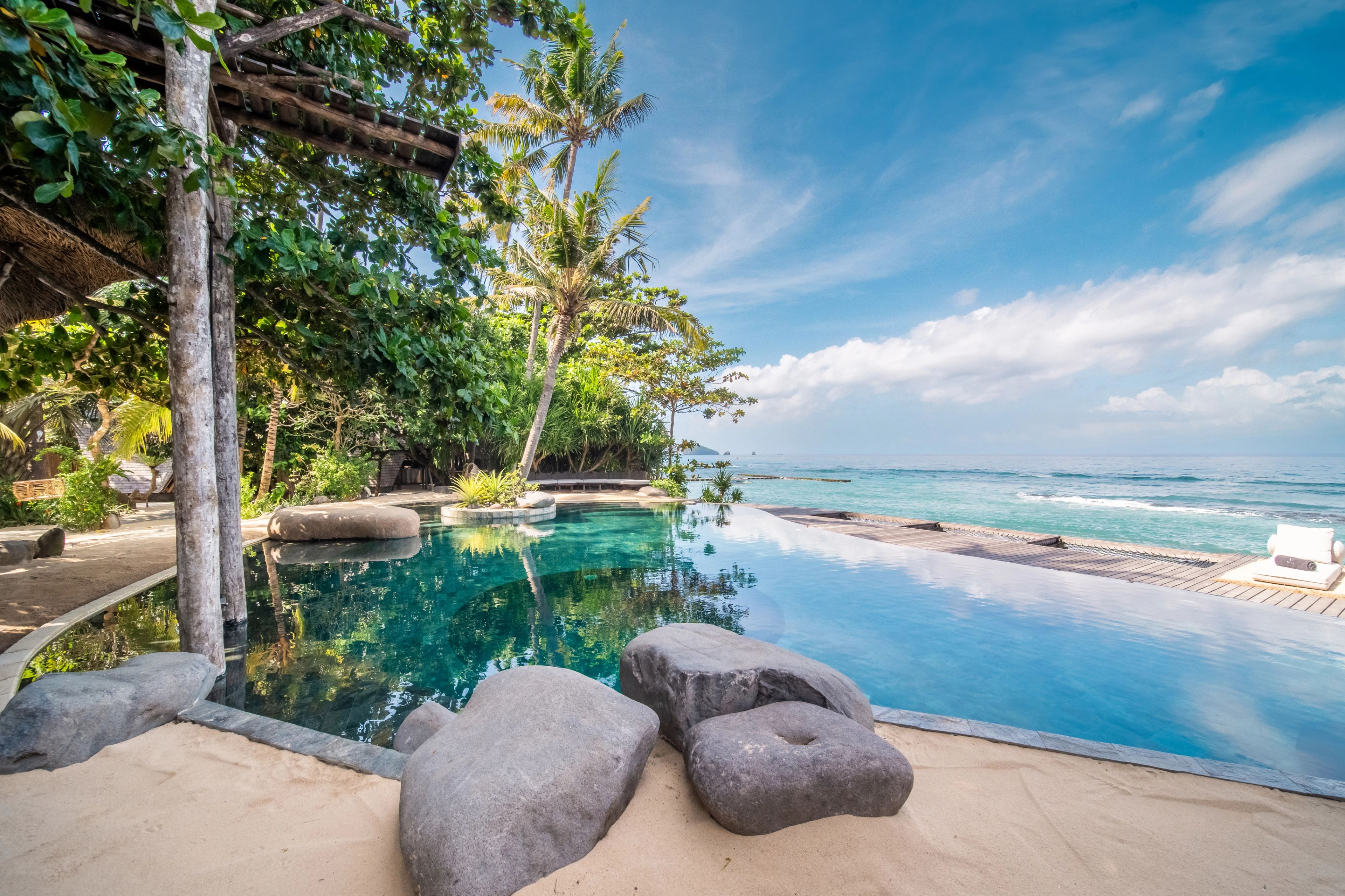 Turquoise pool connected by a curved boardwalk to the beach with palm trees and distant islets at The Seed Bali.