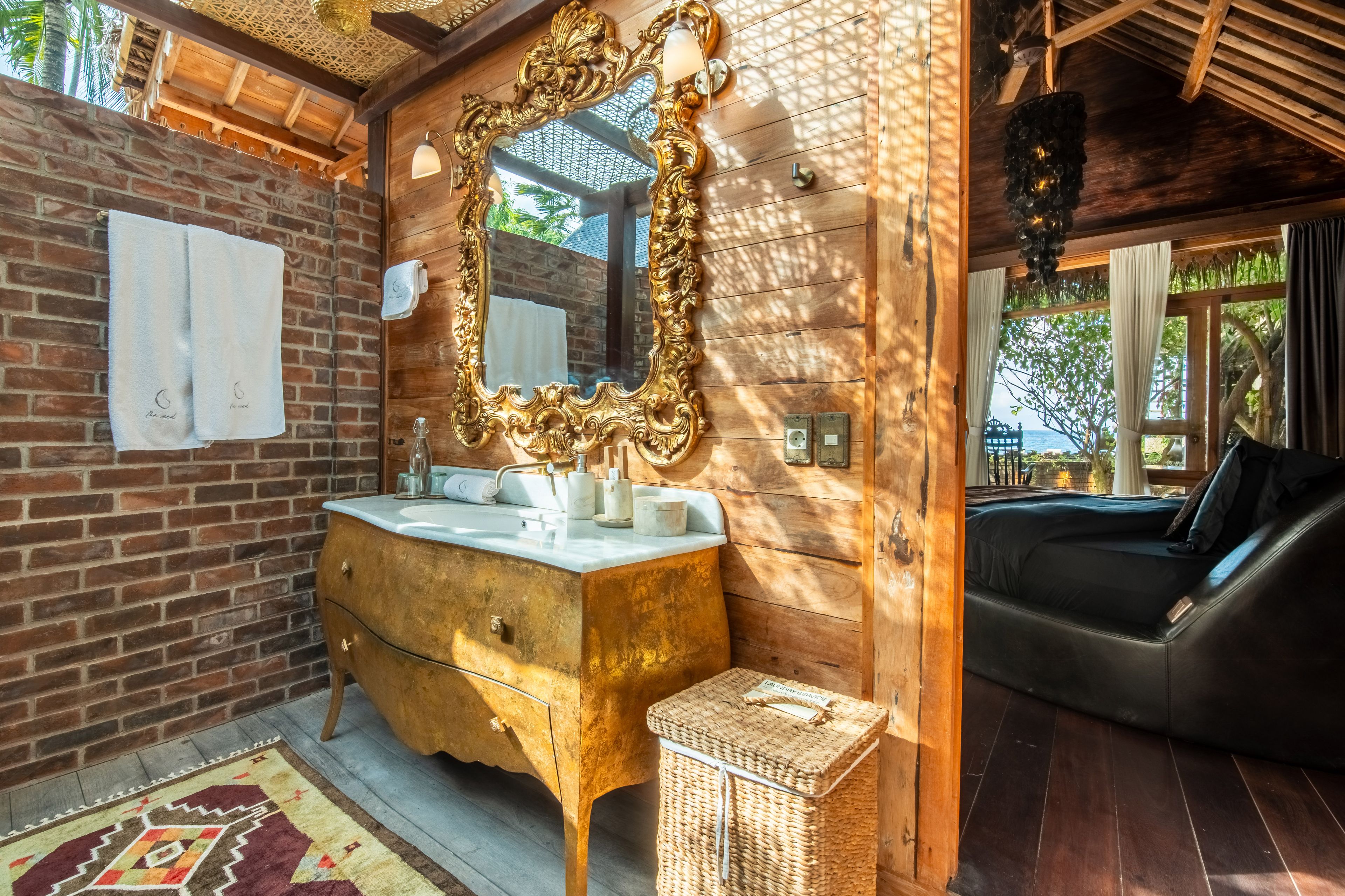 An antique washstand under a gilded mirror against the brick wall, the round bed reflected beyond.