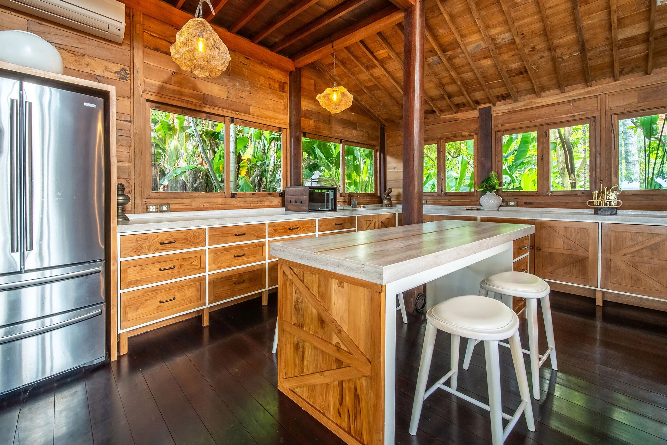 A timber kitchen with an island and stools, windows on two sides looking into the garden.