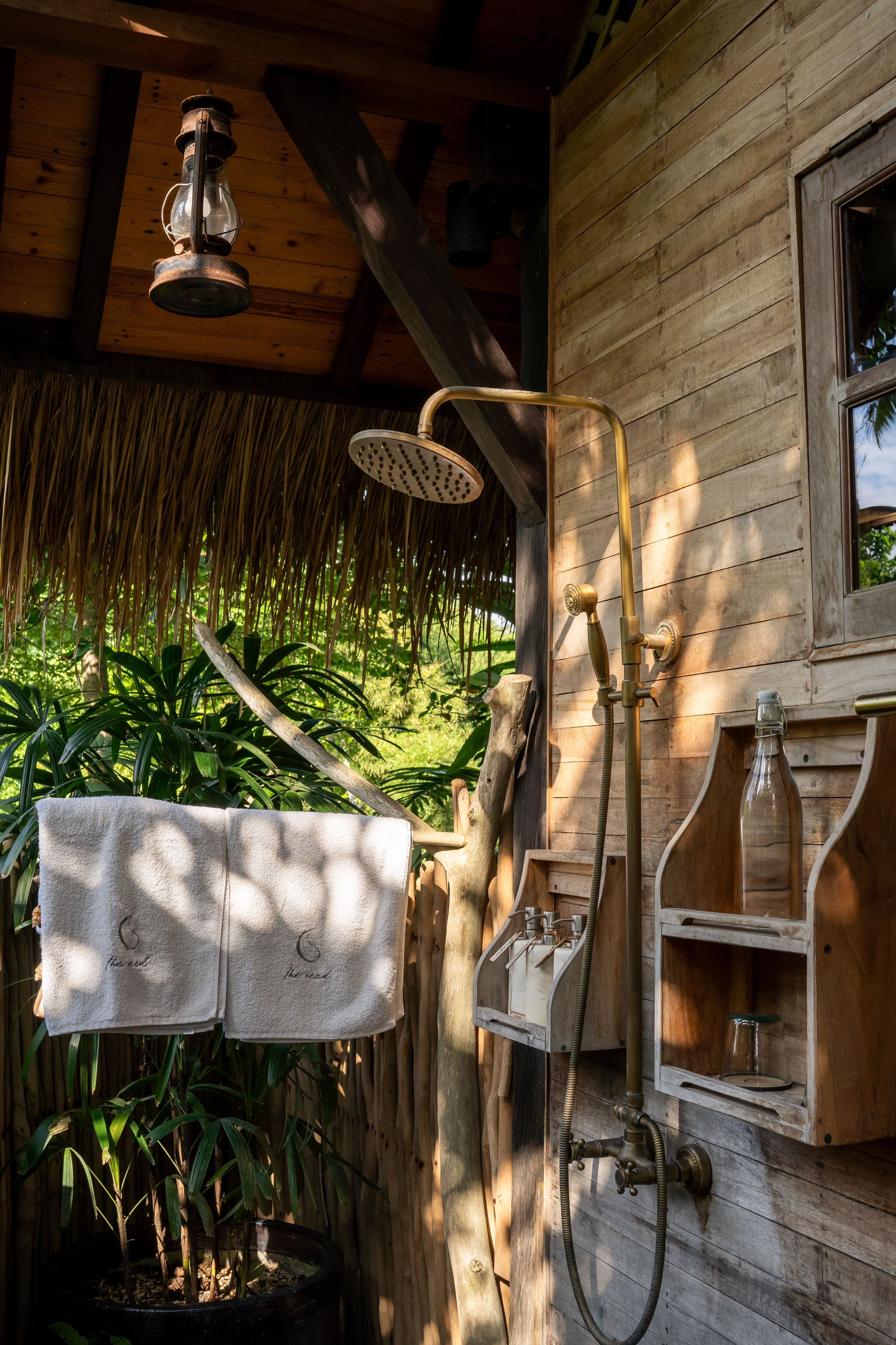 A brass rain shower on the timber wall beside a basin, open to the planting under the thatched eaves.