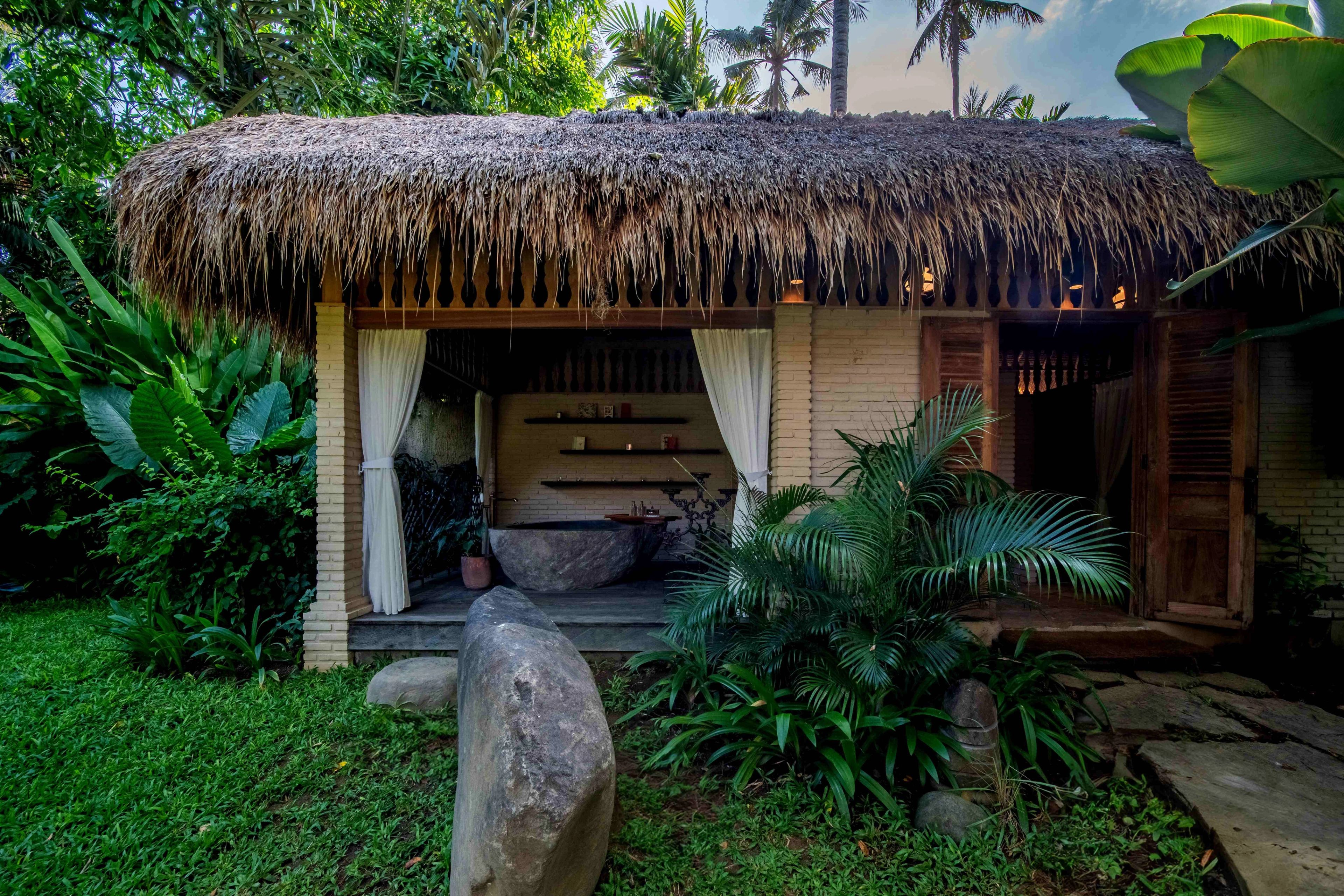 The open-sided thatched bathroom pavilion seen from the lawn, framed by boulders and planting.