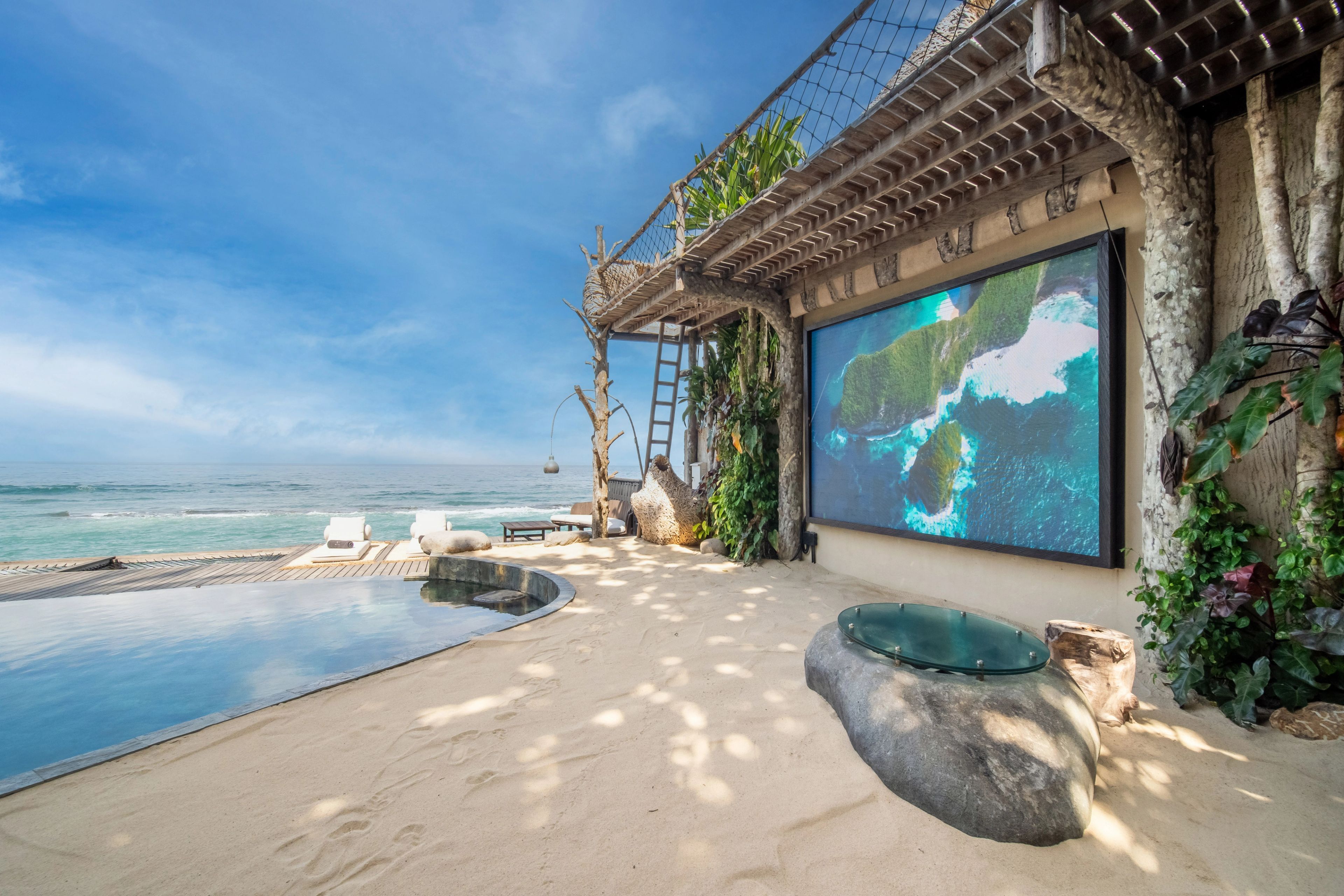 The outdoor screen playing aerial footage above the pool, a stone jacuzzi in the sand beside it.