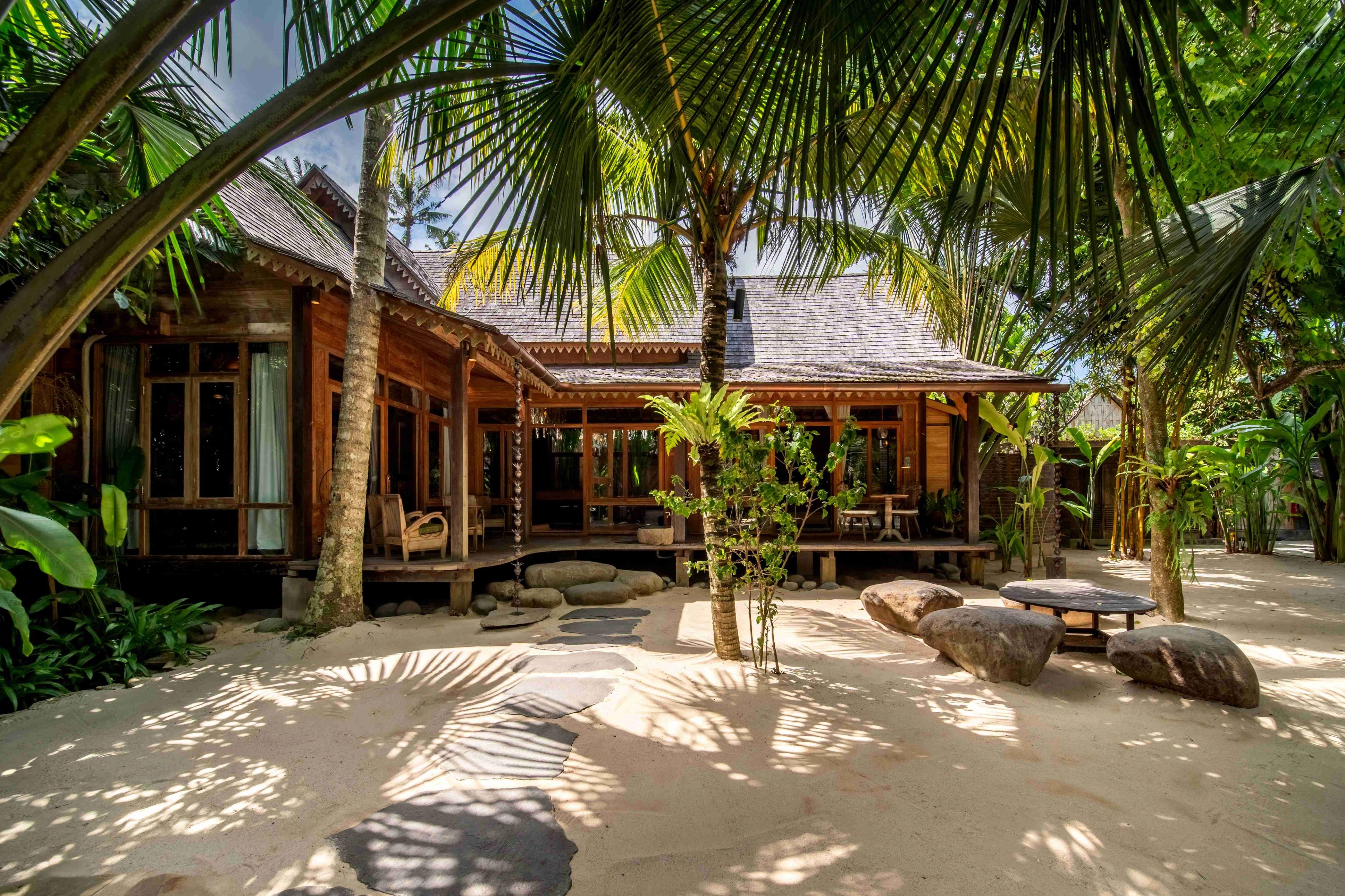 The villa's timber façade across a raked sand courtyard set with boulders, under the palms.