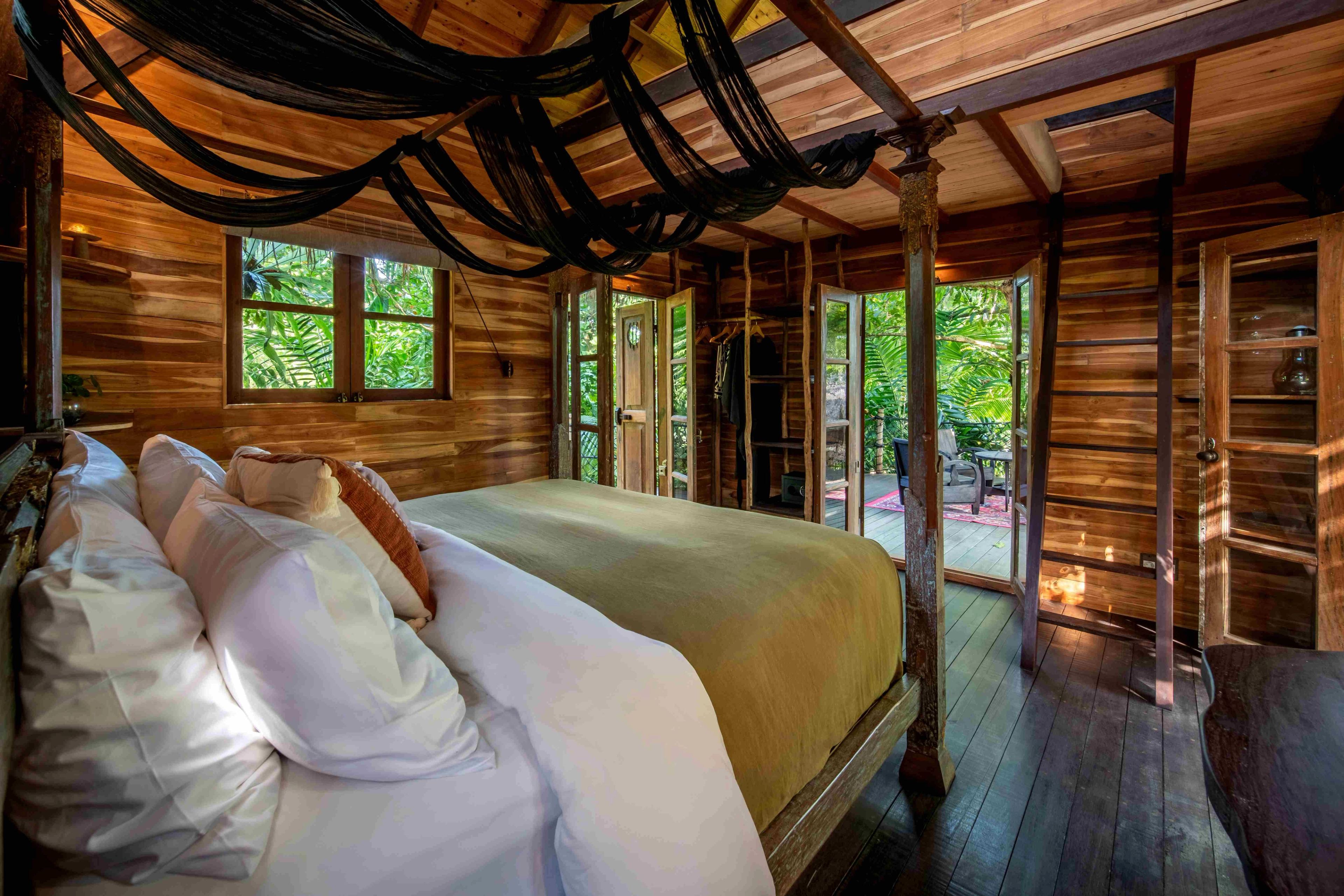 A bed piled with white pillows under a draped canopy, doors open to the deck and the trees.