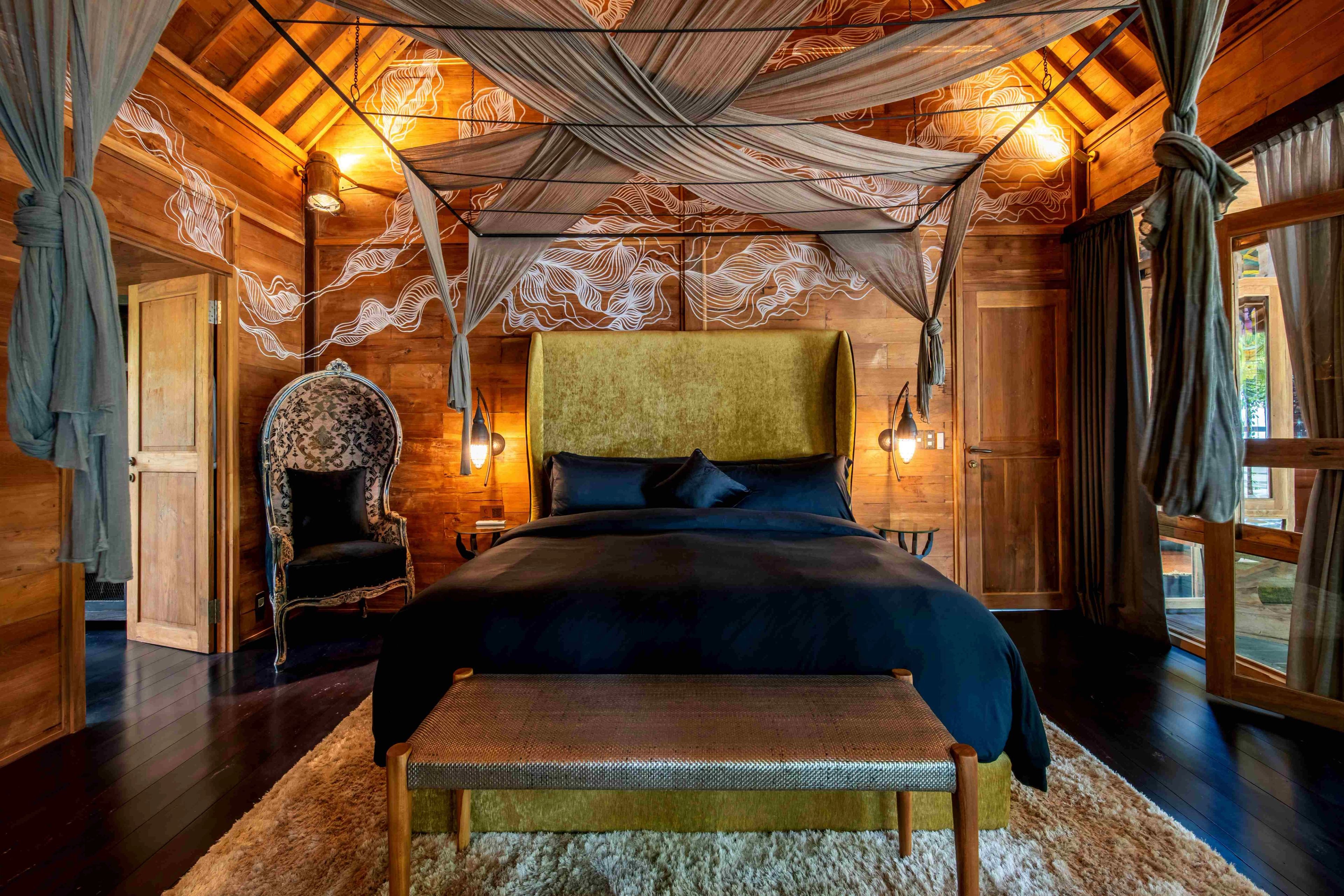 A bed in dark linen beneath a draped mosquito net, a carved chair and bench either side.