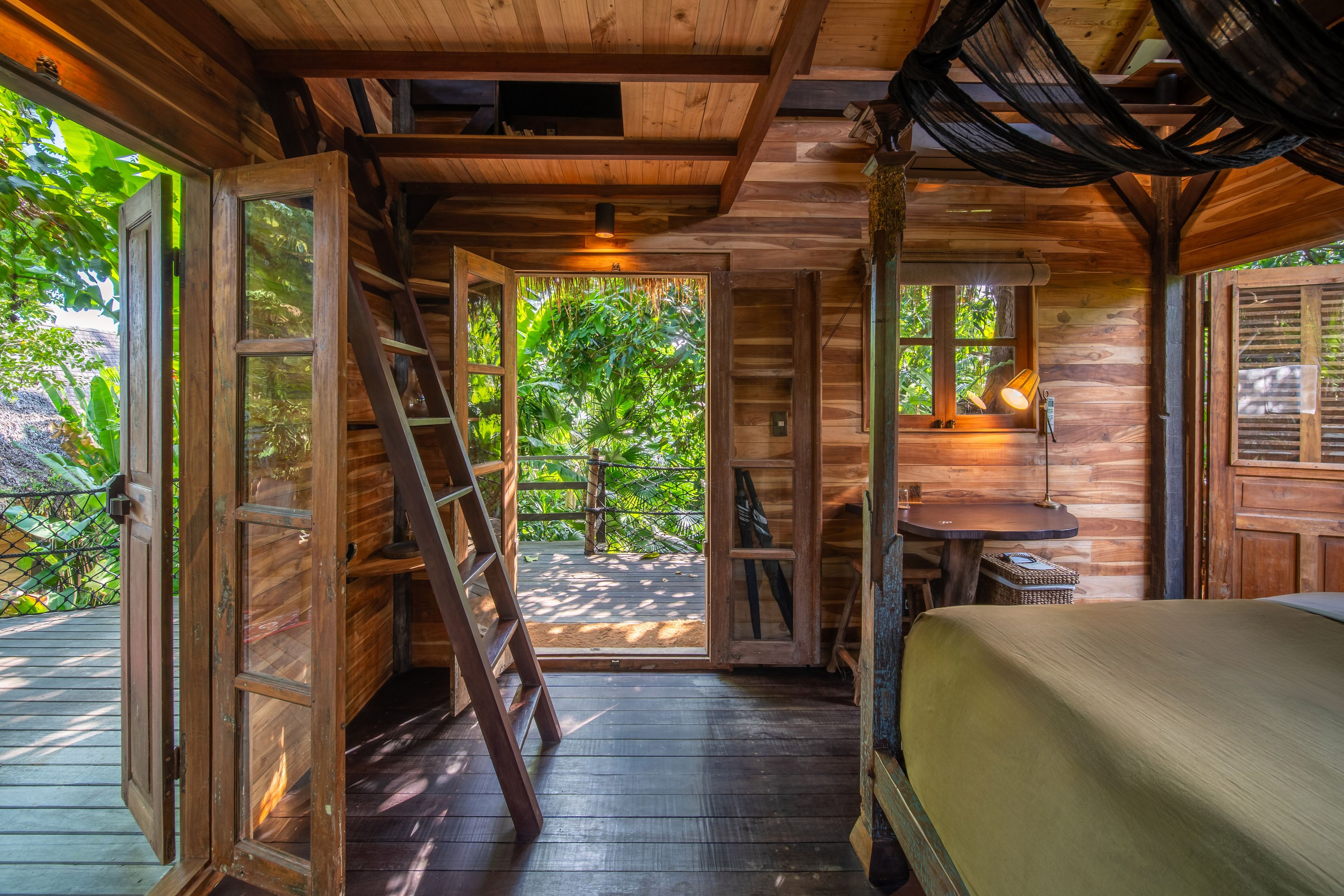 A timber ladder climbing to the loft above the bedroom, with the desk and shuttered window beside it.