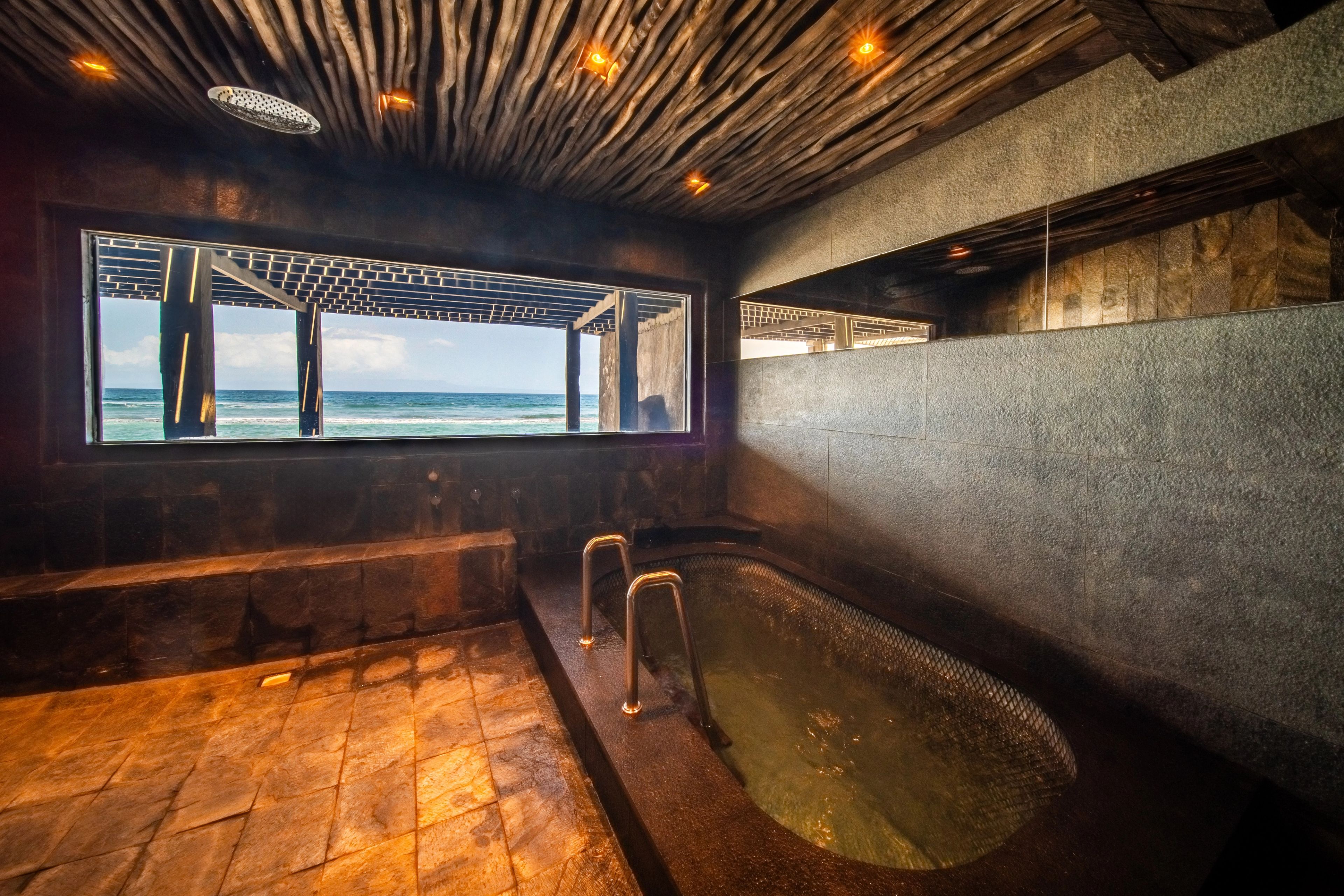 A sunken stone hot tub under a long window framing the sea, with a timber-slat ceiling above.
