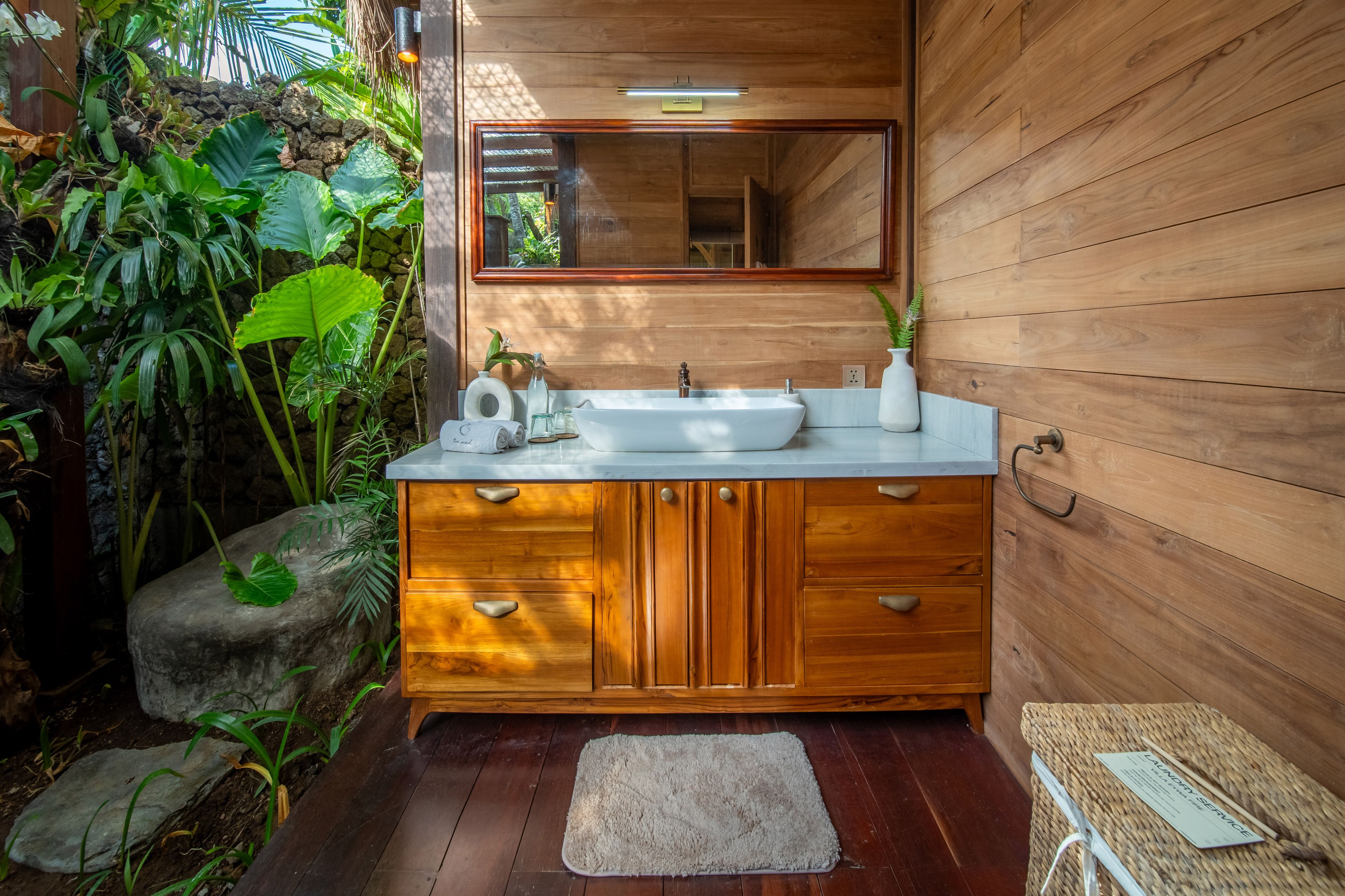 A basin on a timber vanity below a framed mirror, planting growing against the window beside it.