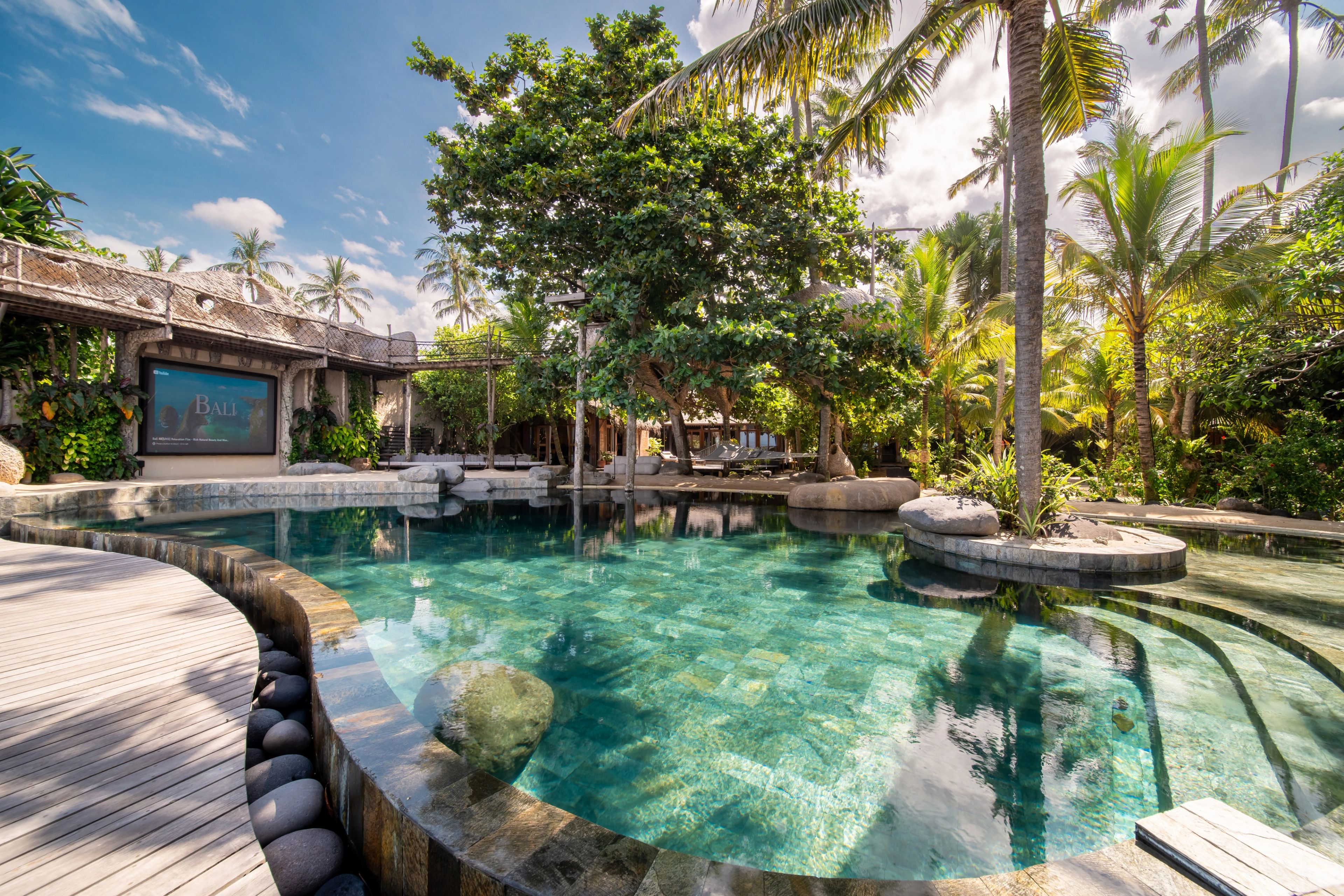 Rounded boulders set along the pool edge beside the timber deck, with palms and the pavilion behind.