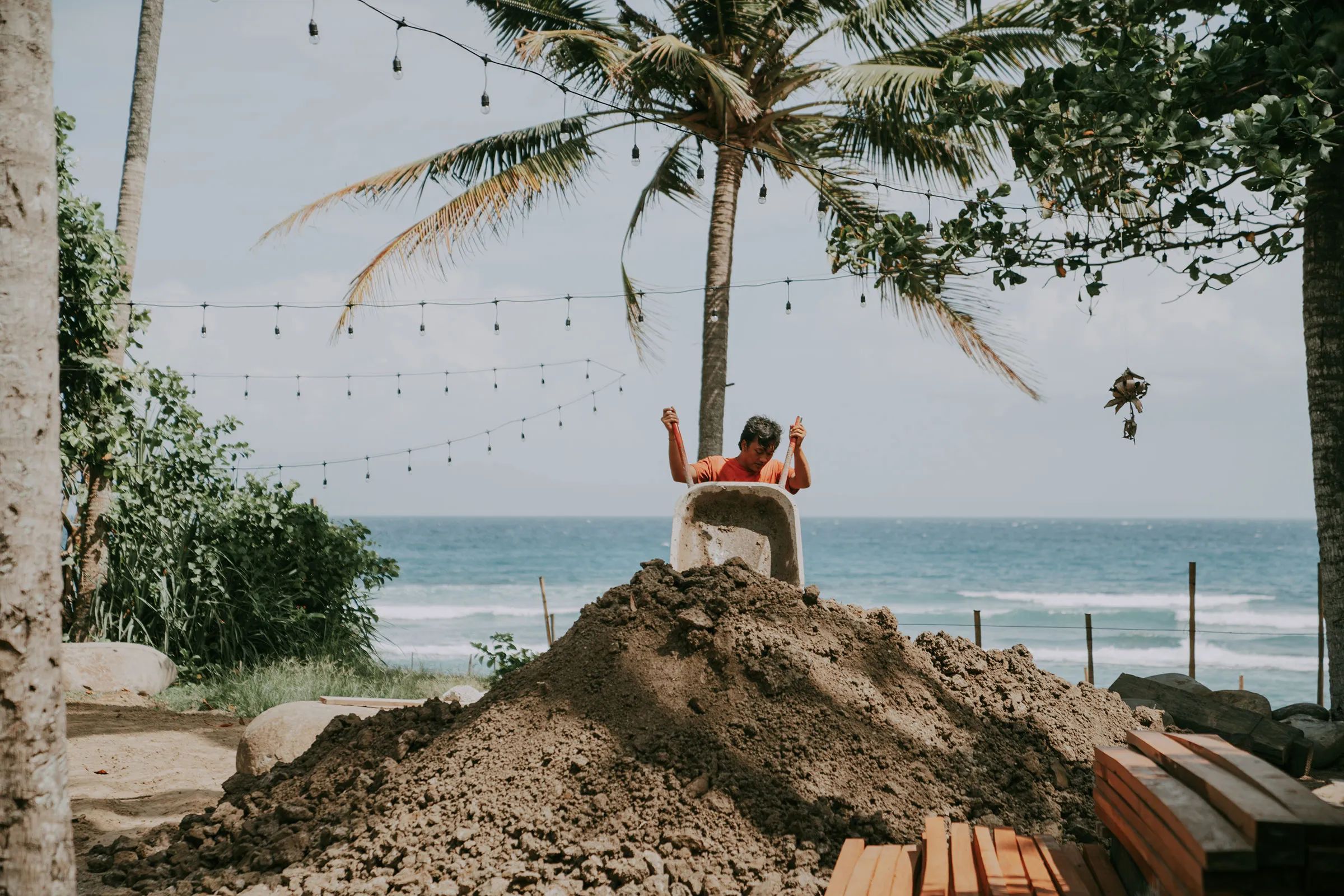 A builder tipping a wheelbarrow of earth onto a mound during construction at The Seed, with stacked timber, palms and the sea behind