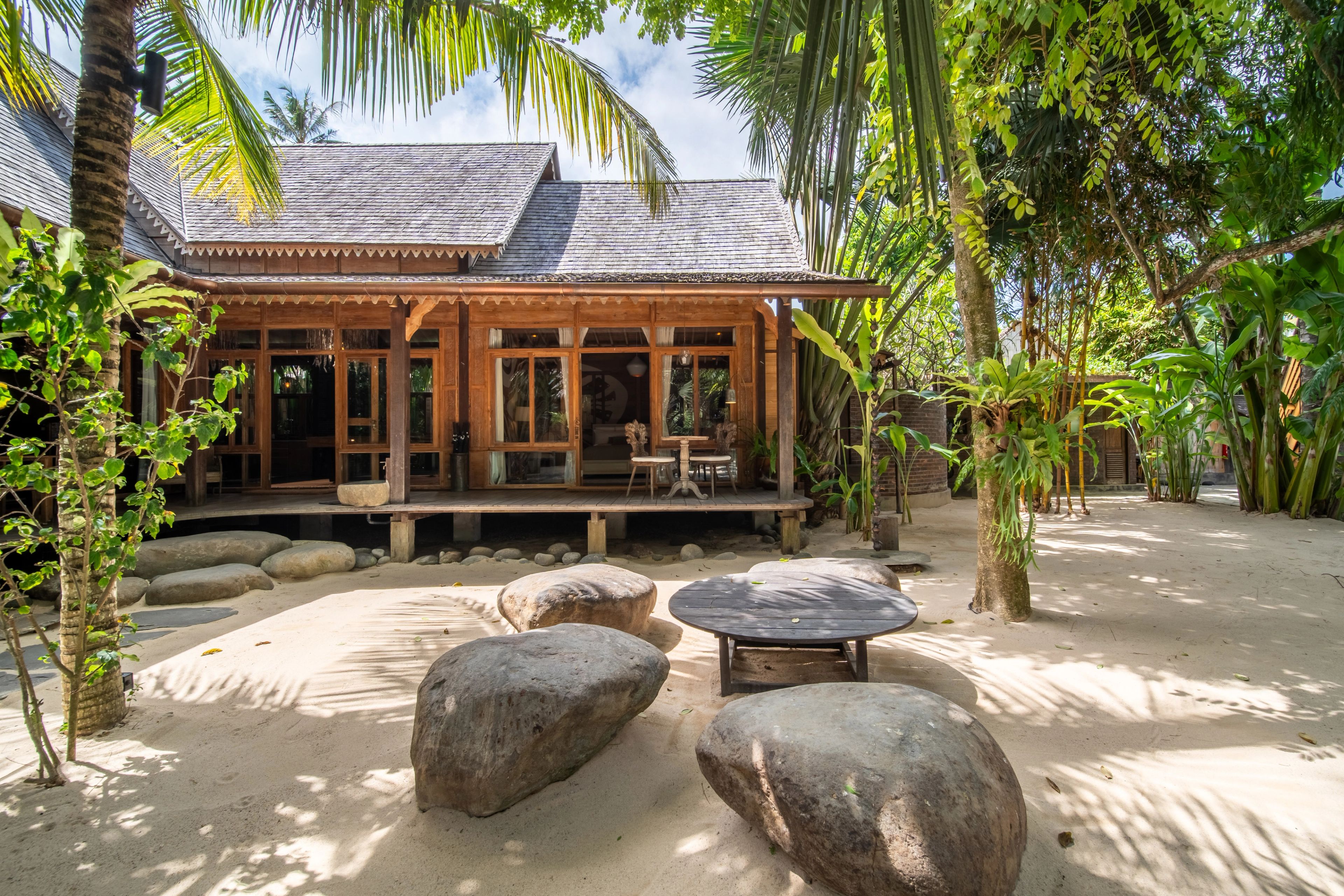 The villa's veranda across the sand courtyard, boulders and a low table set out on the sand.