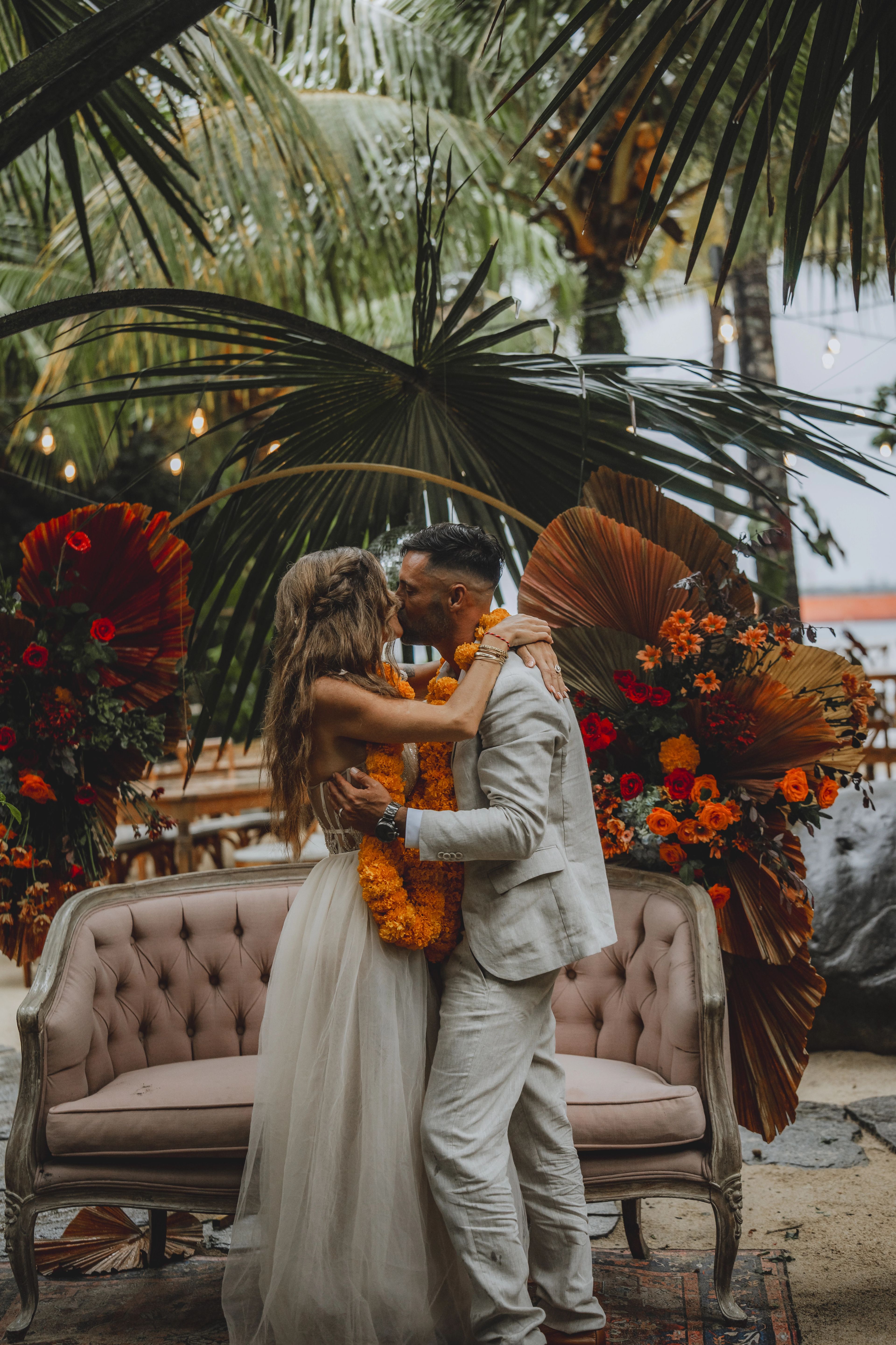 A couple kissing on a tufted sofa, surrounded by marigold, red and dried palm arrangements.