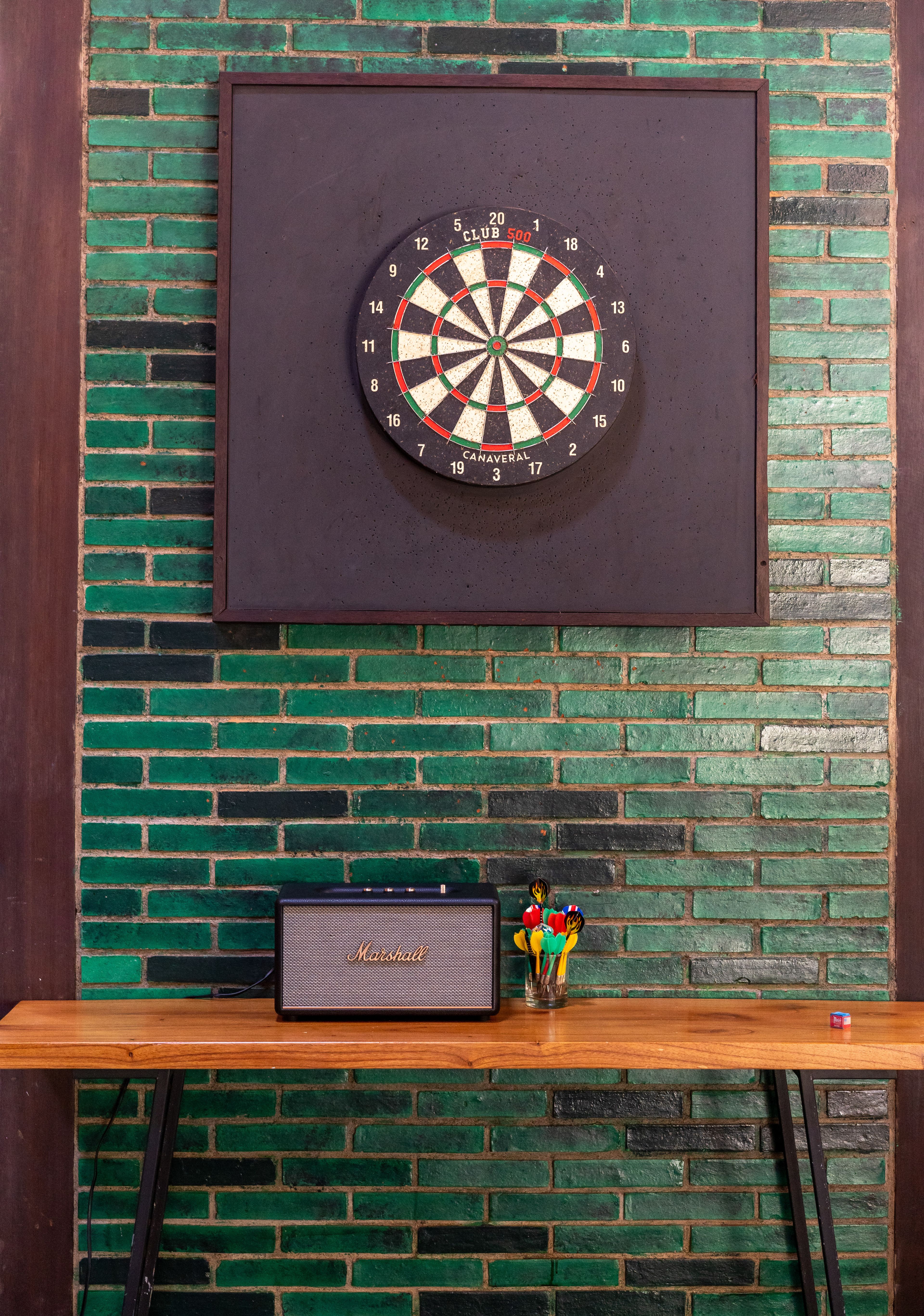 A dartboard mounted on the green brick wall above a Marshall amplifier.