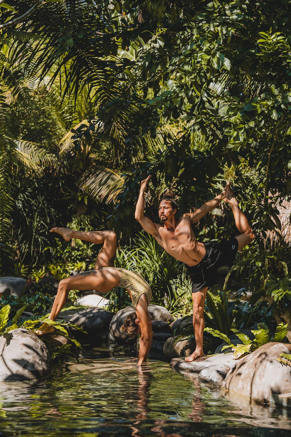 A man moving through a low, twisting pose on the grass, surrounded by dense planting.