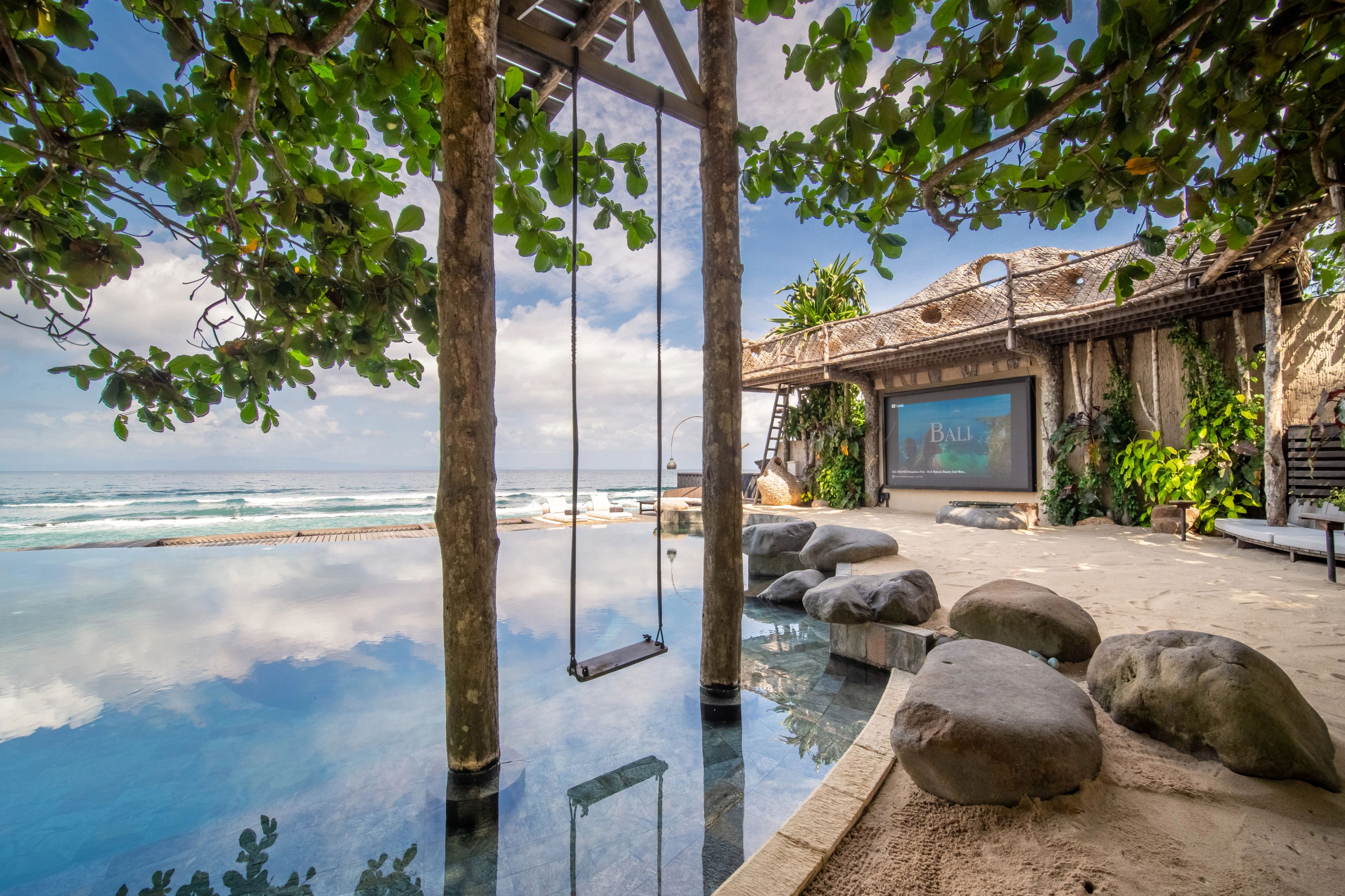 A timber swing hanging from a tree over the pool, the cinema screen set into the wall beyond.