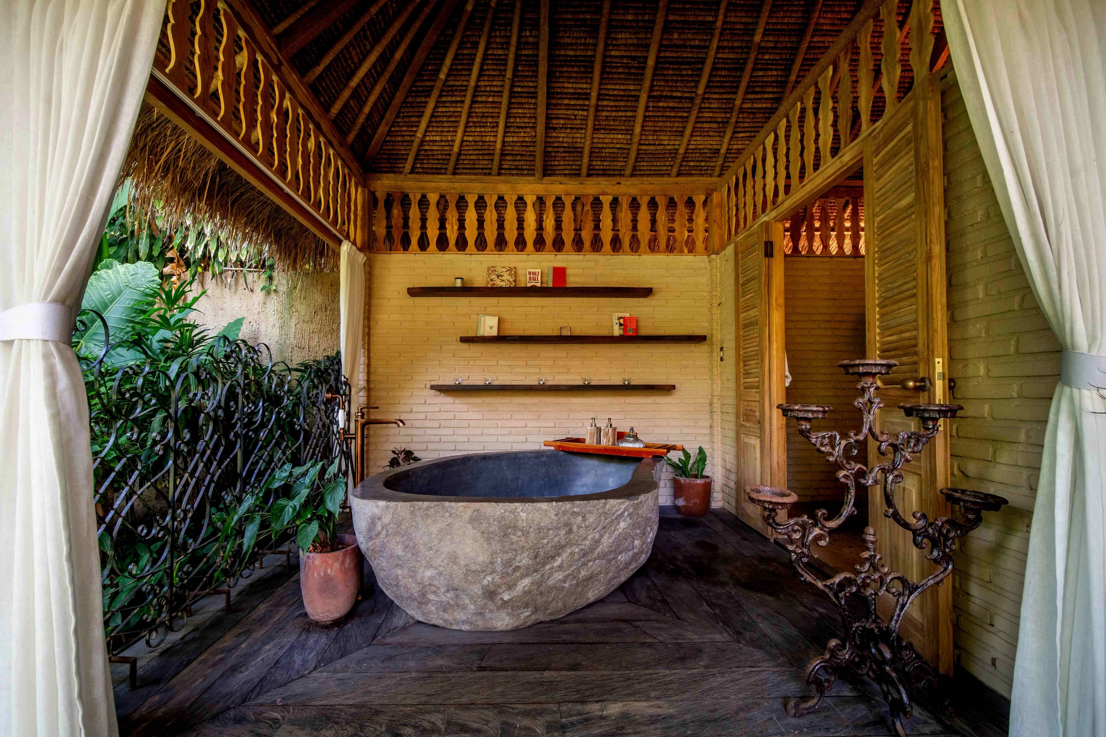 A carved stone bathtub on a dark timber floor between a planted wall and a curtained doorway.