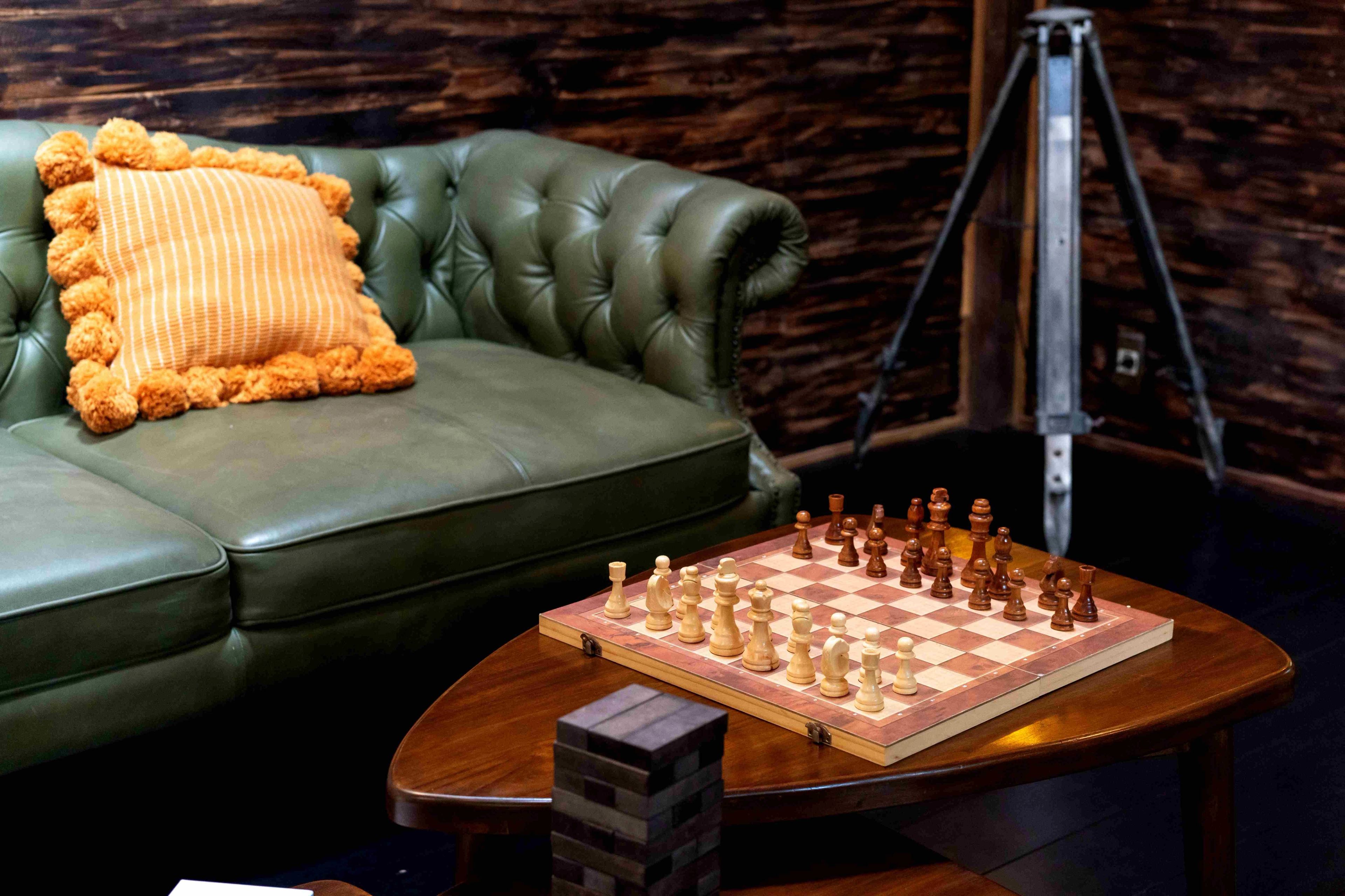 A chess set laid out on a curved timber table beside a green leather sofa.