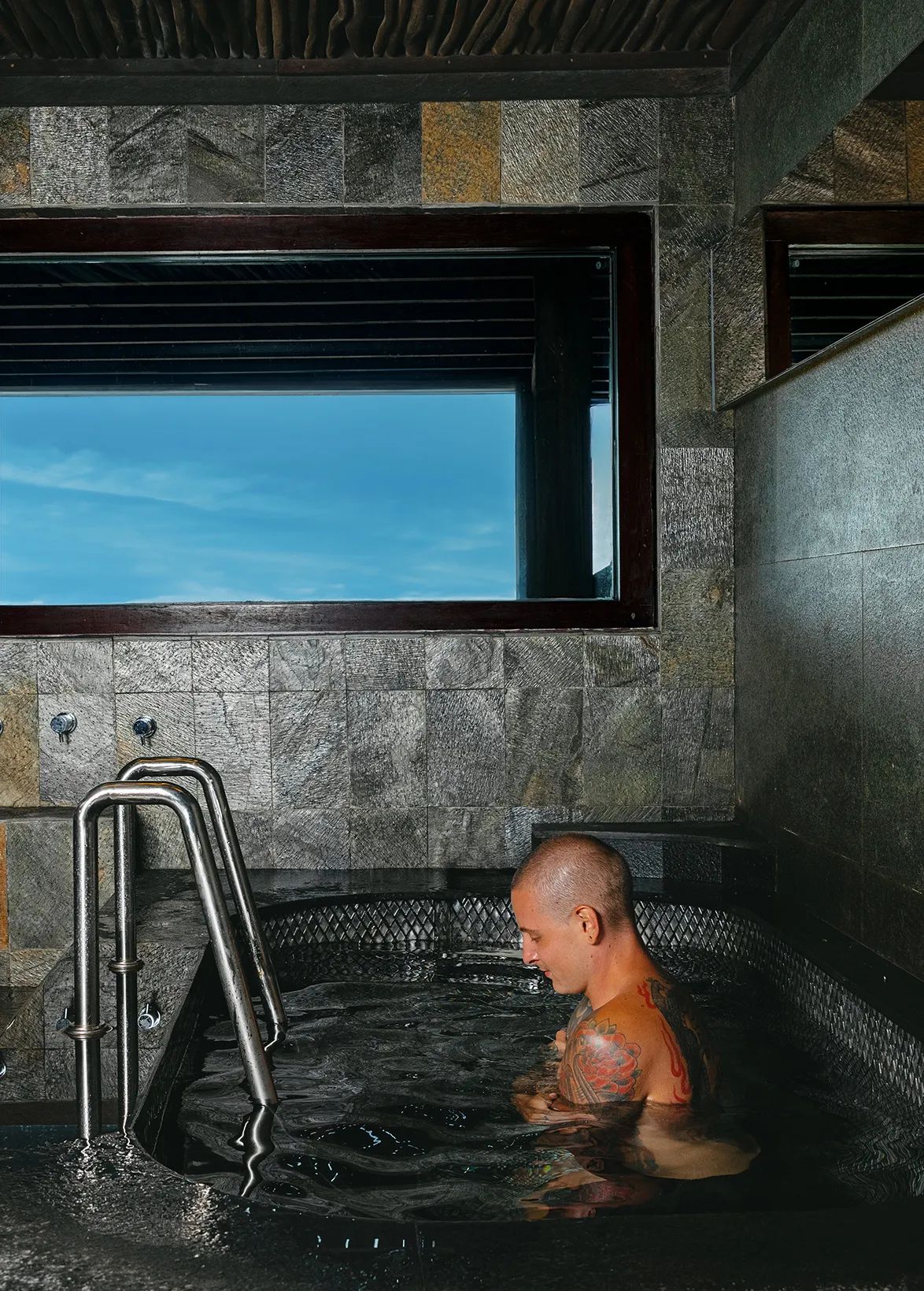 A guest in the cold plunge with the handrail beside him, the sky visible through the window above.