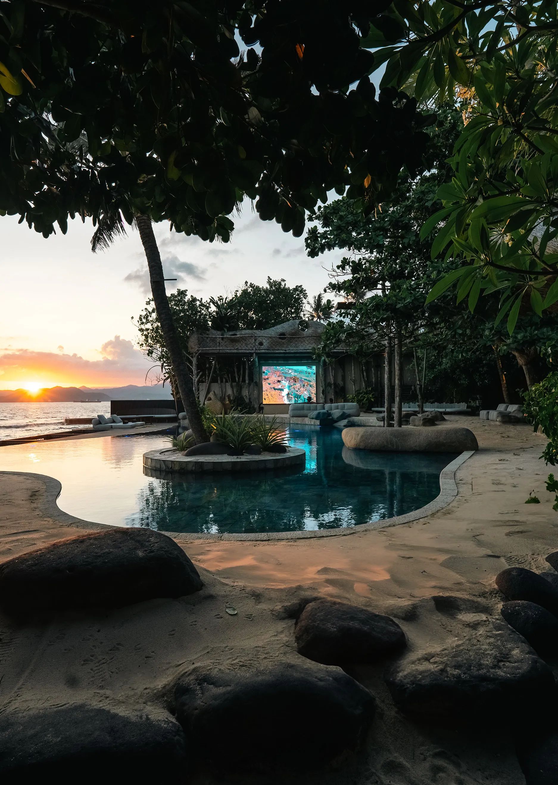 The outdoor screen lit beside the pool as the sun sets over the water behind the palms.