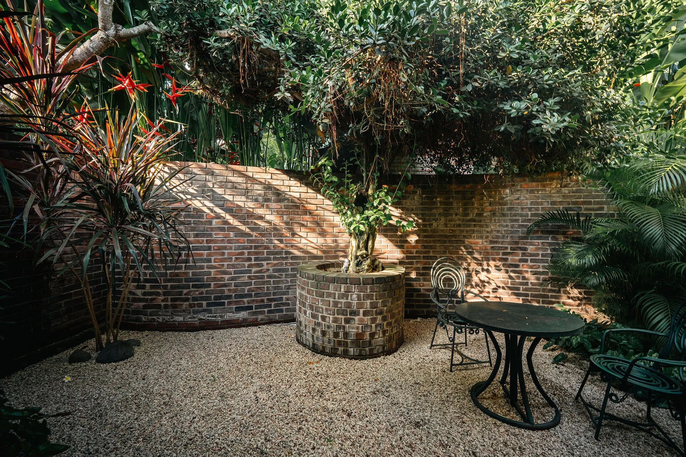 A curved brick wall enclosing a gravel courtyard with a bistro table and a tree at its centre.