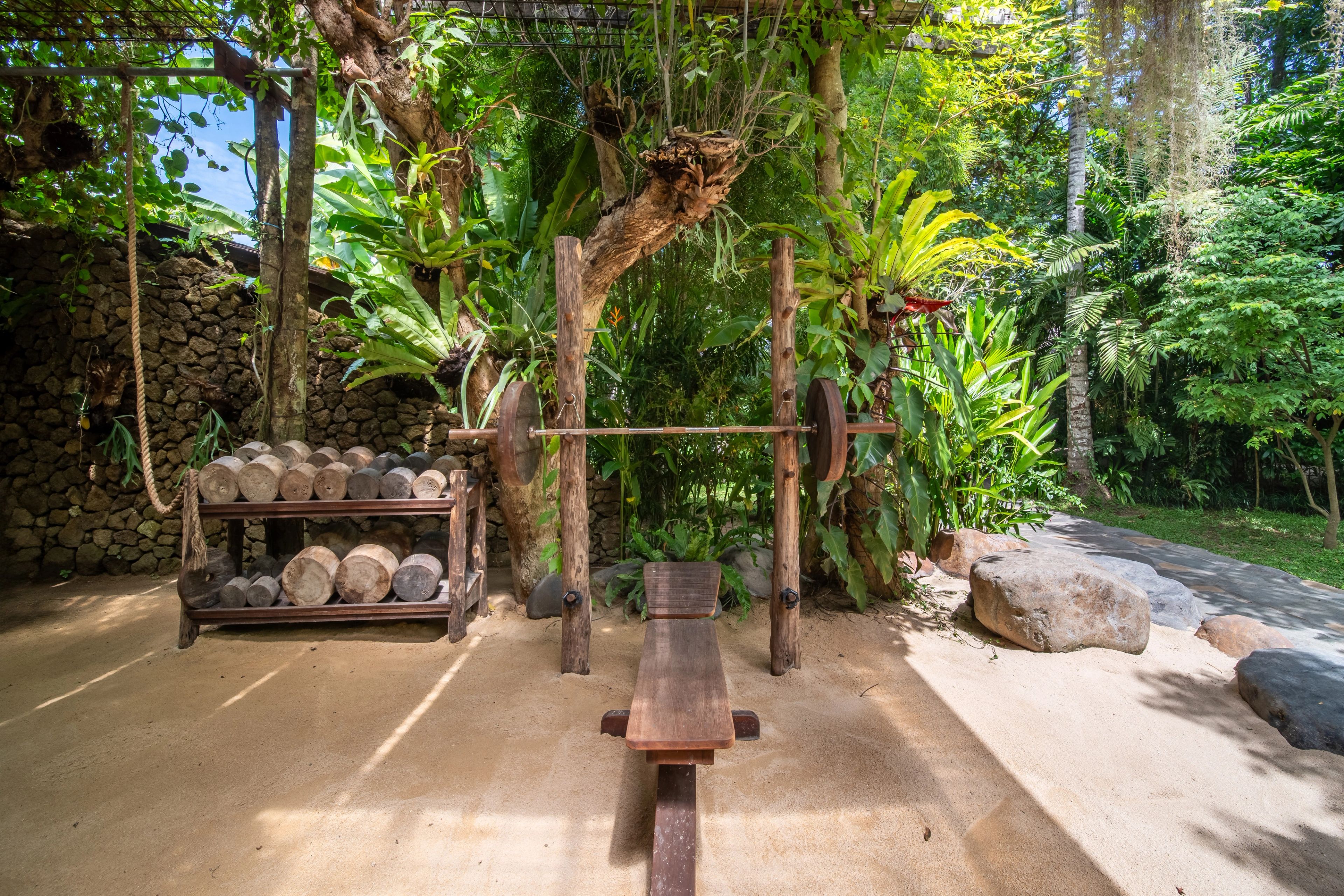 A timber squat rack and bench on the sand floor, backed by the stone wall and dense planting.