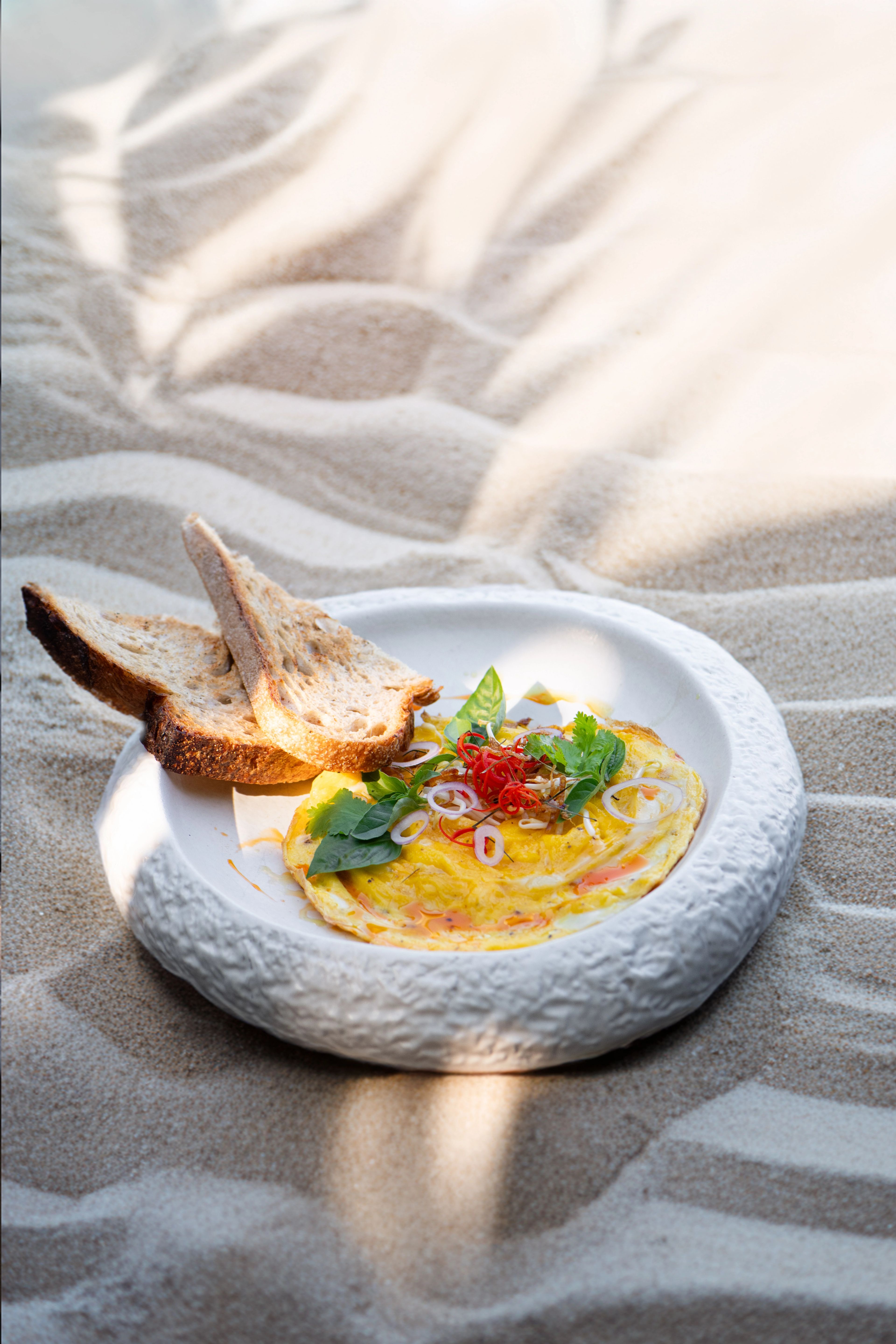 An omelette with chilli and basil, served with toasted sourdough on a stoneware plate set on the sand.