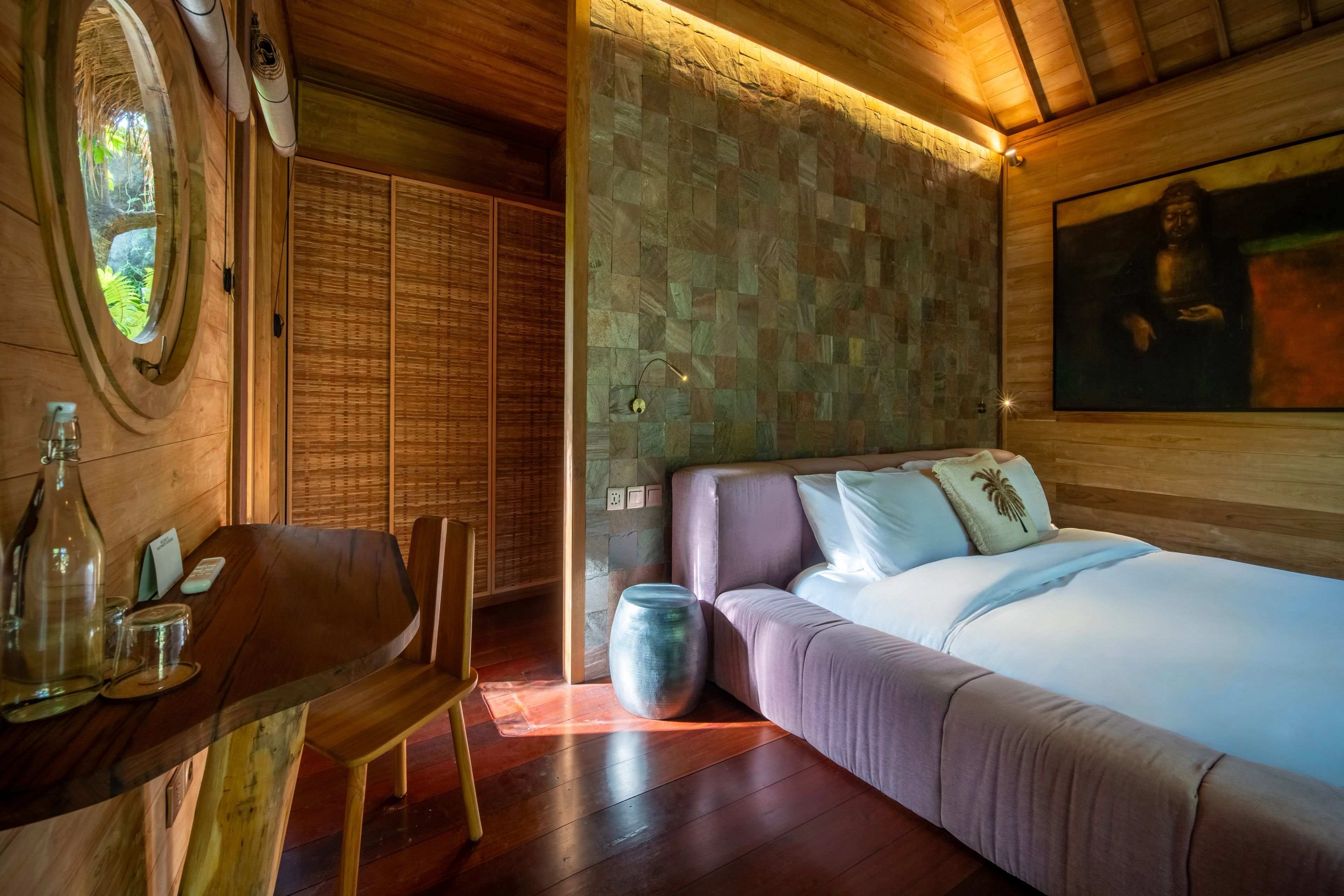 A pink upholstered bed beside a timber desk, a framed painting on the tiled wall behind.