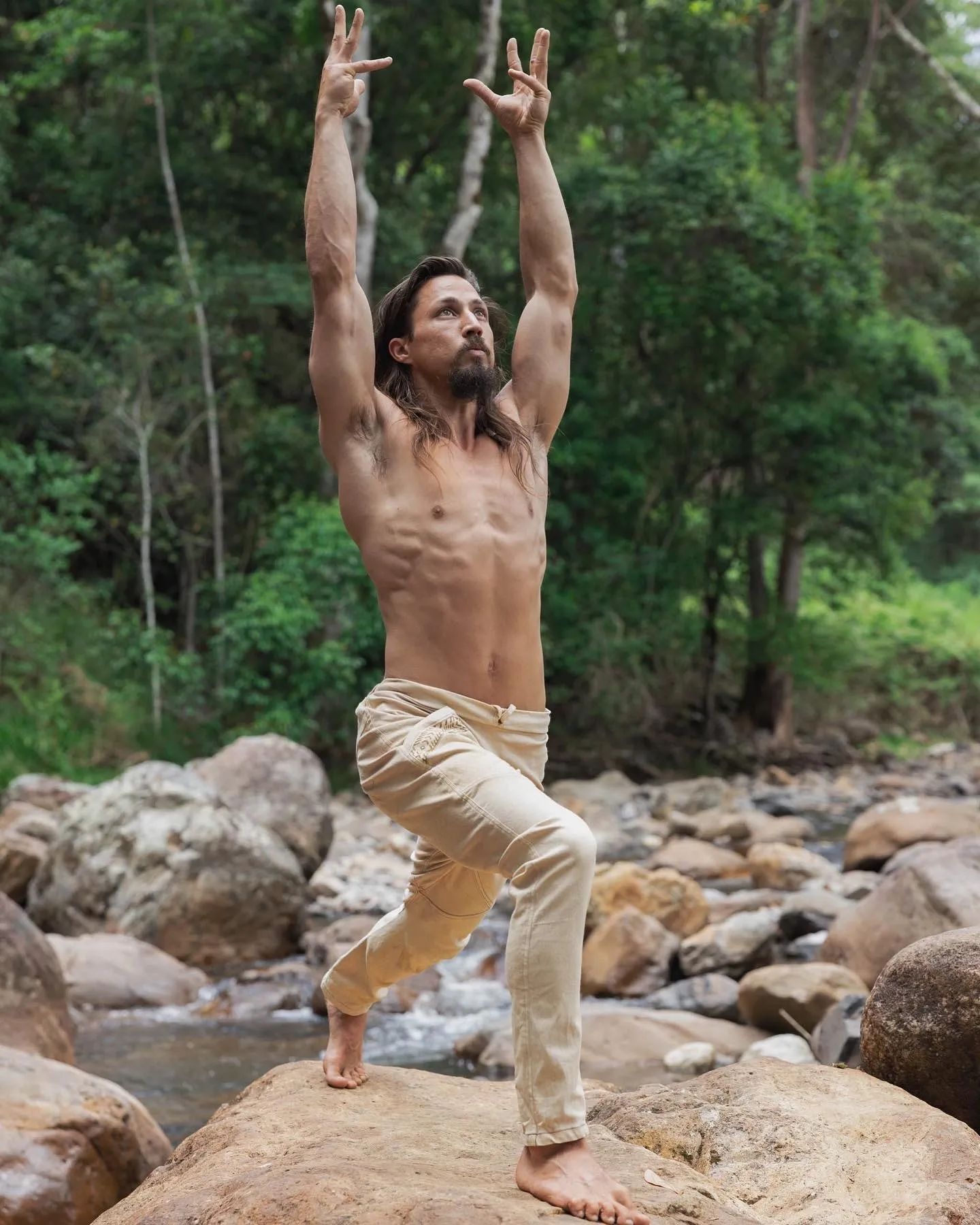 A man stretching upward on the river boulders with the jungle behind him.