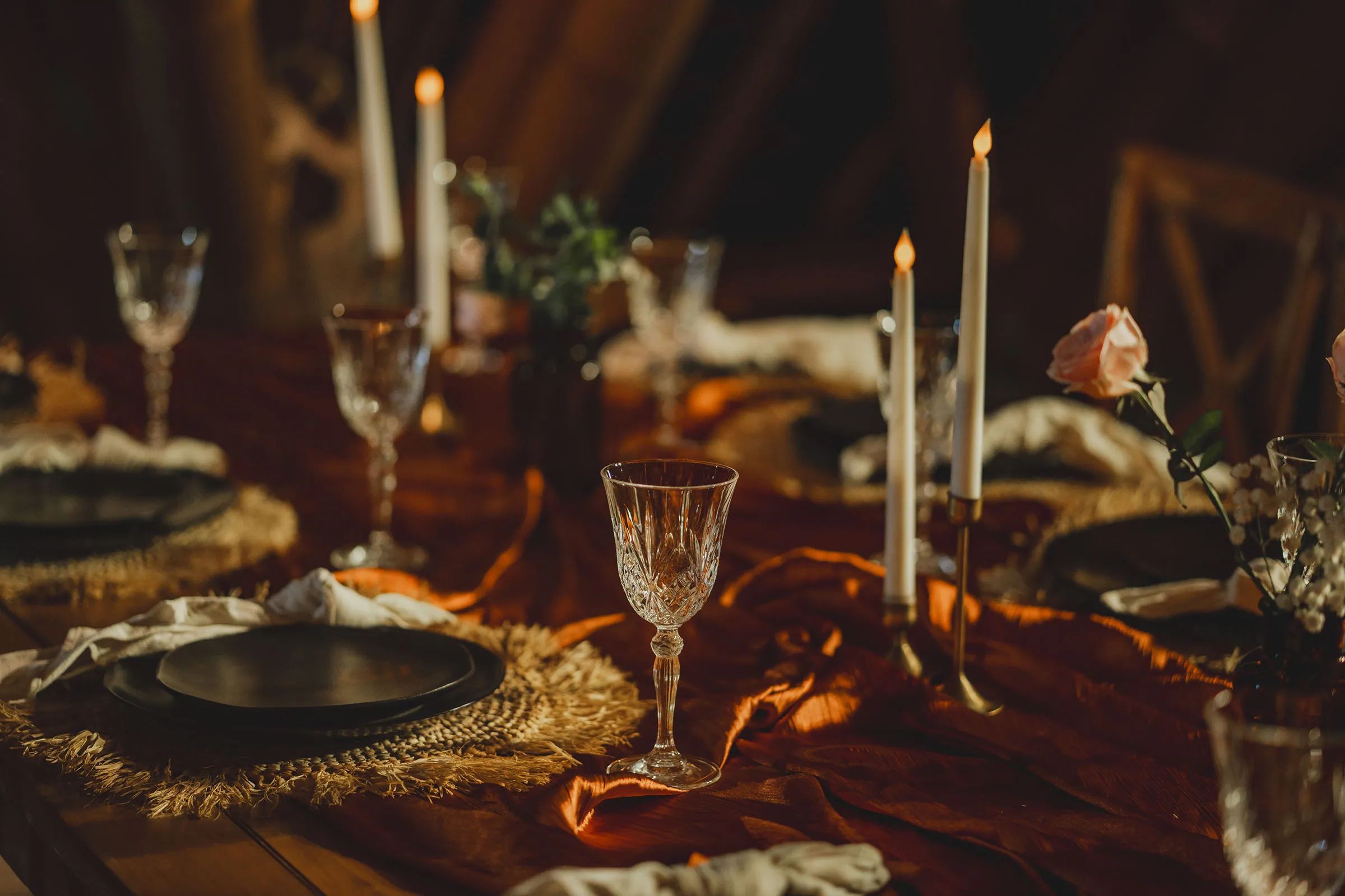 Cut-glass goblets, tapers and a rose along a rust-coloured runner on a table laid for dinner.
