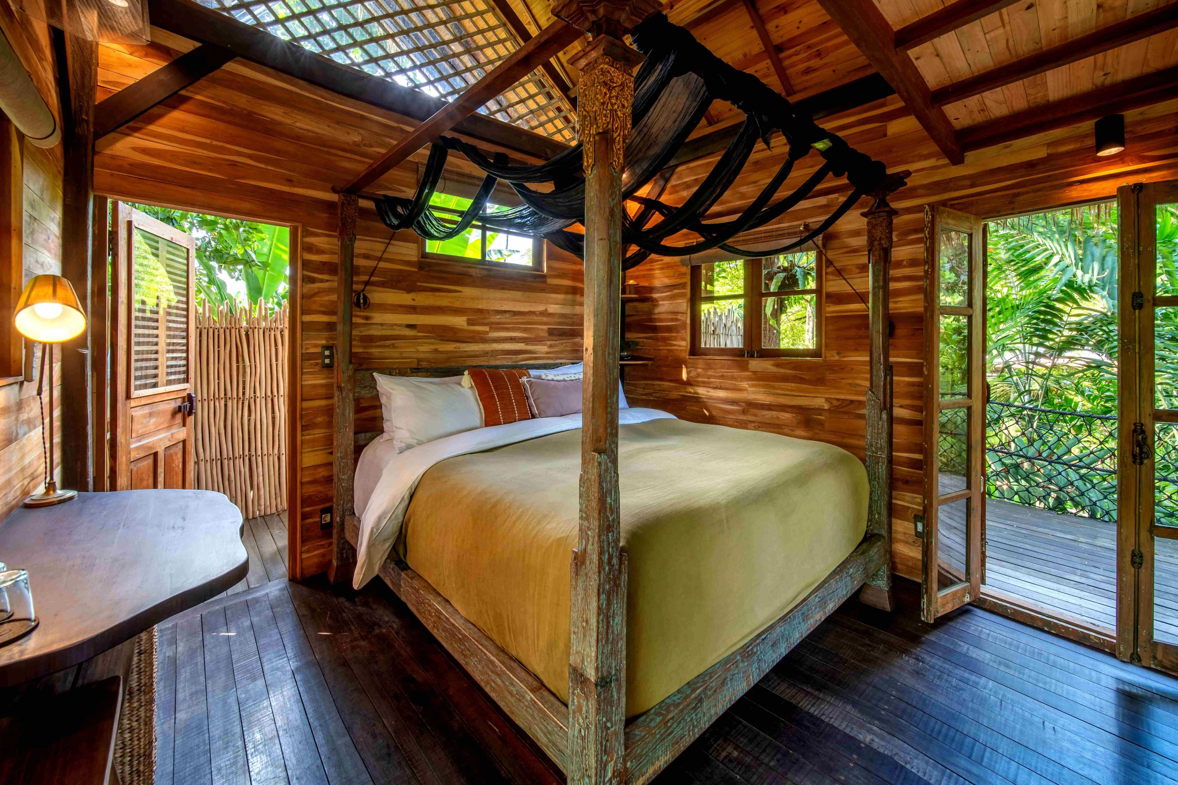 A four-poster bed with its netting tied back, under the timber roof with doors open on both sides.
